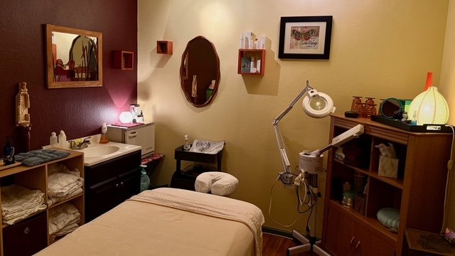 A massage or treatment room with a massage table, shelves with towels, a mirror, and various bottles and decorative items.