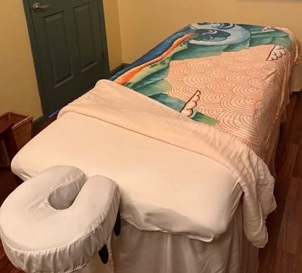 A massage table with a colorful blanket and a white face cradle, situated in a room with cream walls and a closed dark green door.
