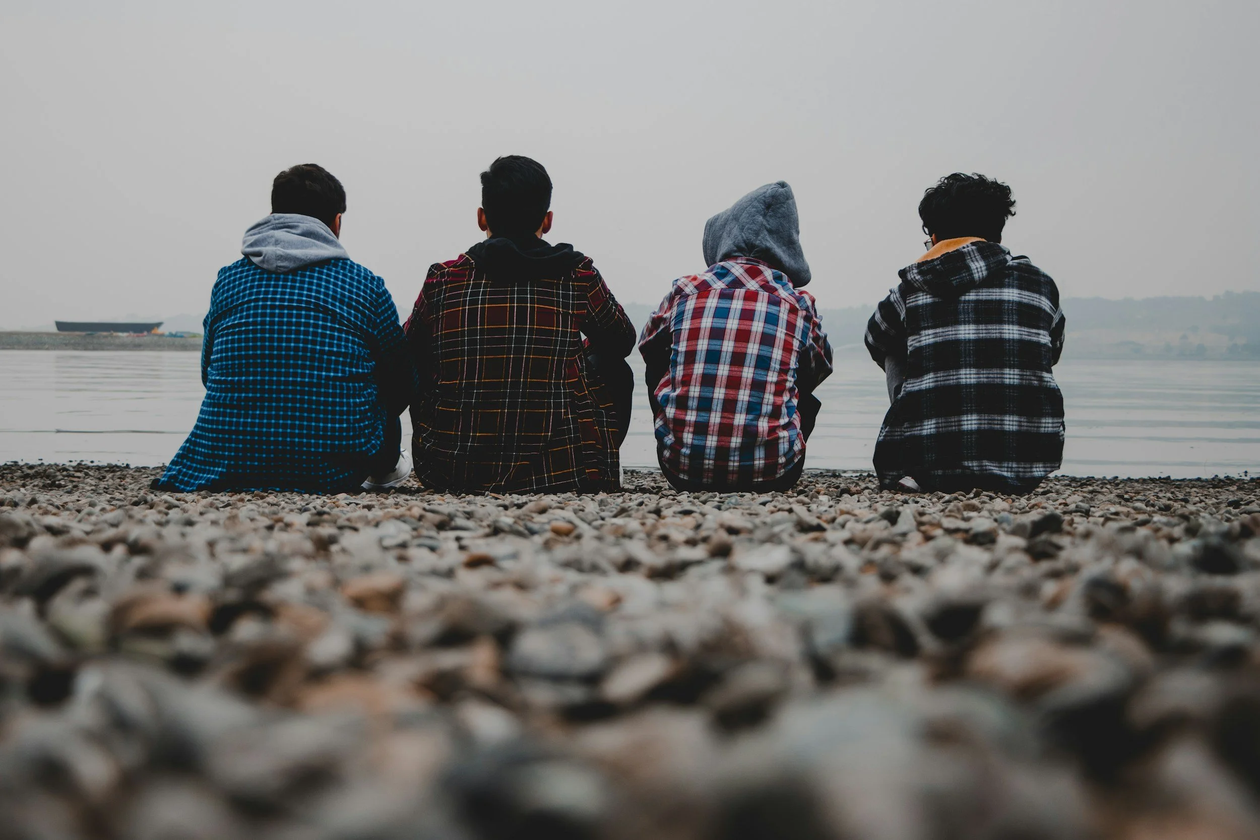 Four young men sitting on a pebbled beach, facing a calm body of water, with a distant ship visible on the horizon, wearing casual hoodies and jackets.