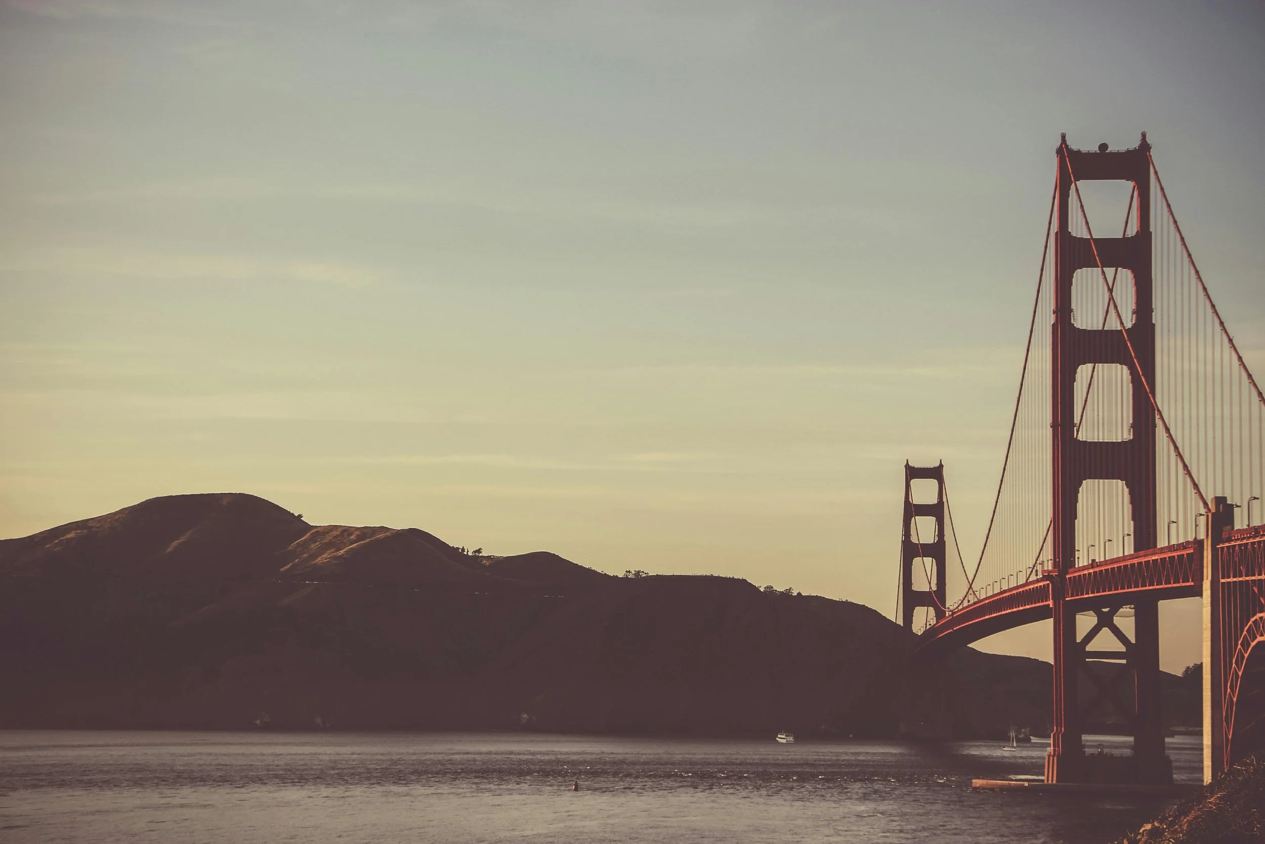 The Golden Gate Bridge with hills in the background and water in the foreground, captured during sunset or sunrise.