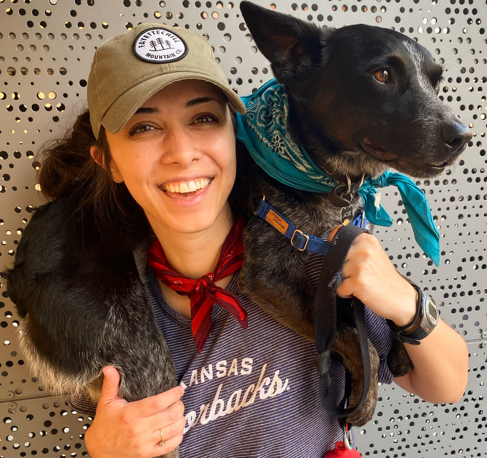 A woman smiling and wearing a tan cap and a red bandana around her neck, is carrying a black dog with brown eyes on her shoulders. The dog has a blue bandana and a harness, and looks to the side. The background is a perforated metallic surface.
