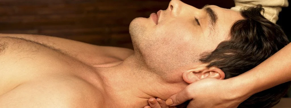 A shirtless man lying down with his eyes closed while a hand touches his neck.