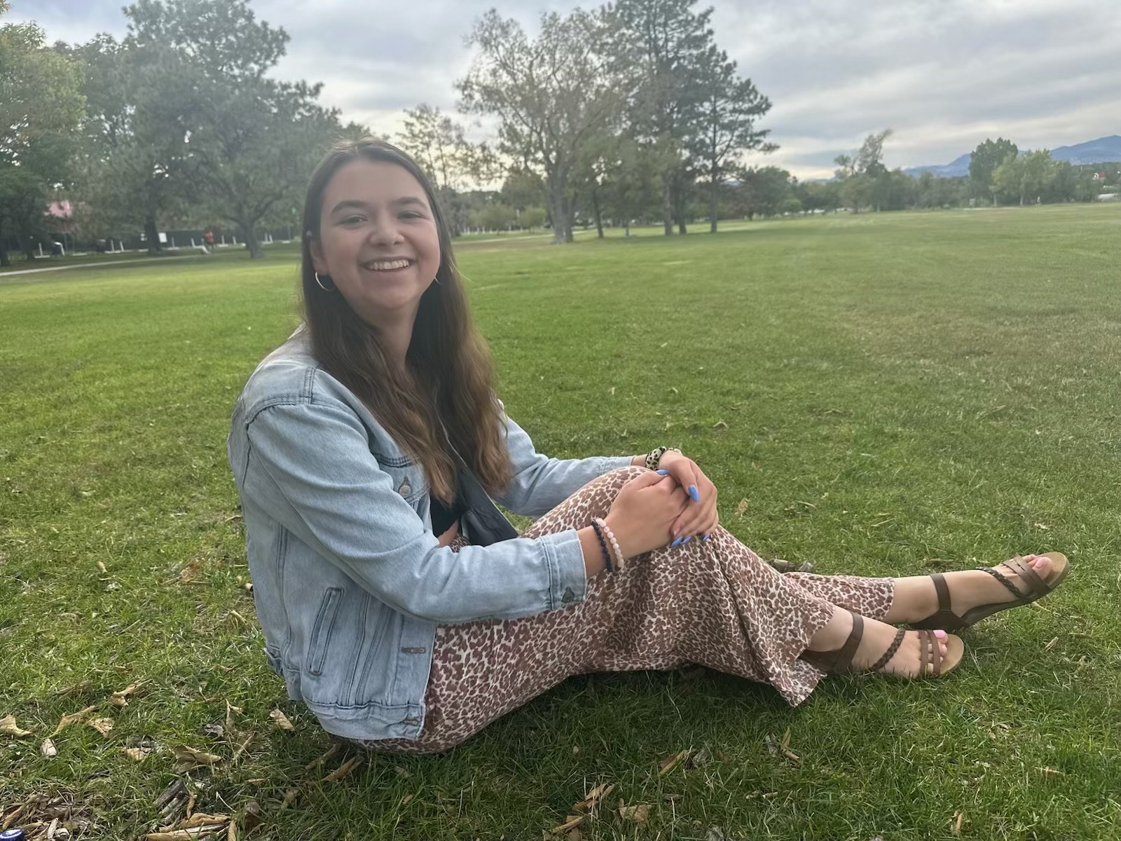 Smiling young woman sitting on grass in a park during daytime, wearing a denim jacket, leopard print pants, and sandals, with trees and cloudy sky in the background.