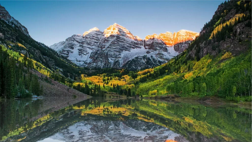 Snow-capped mountains behind a forested valley and a calm lake that reflects the landscape.