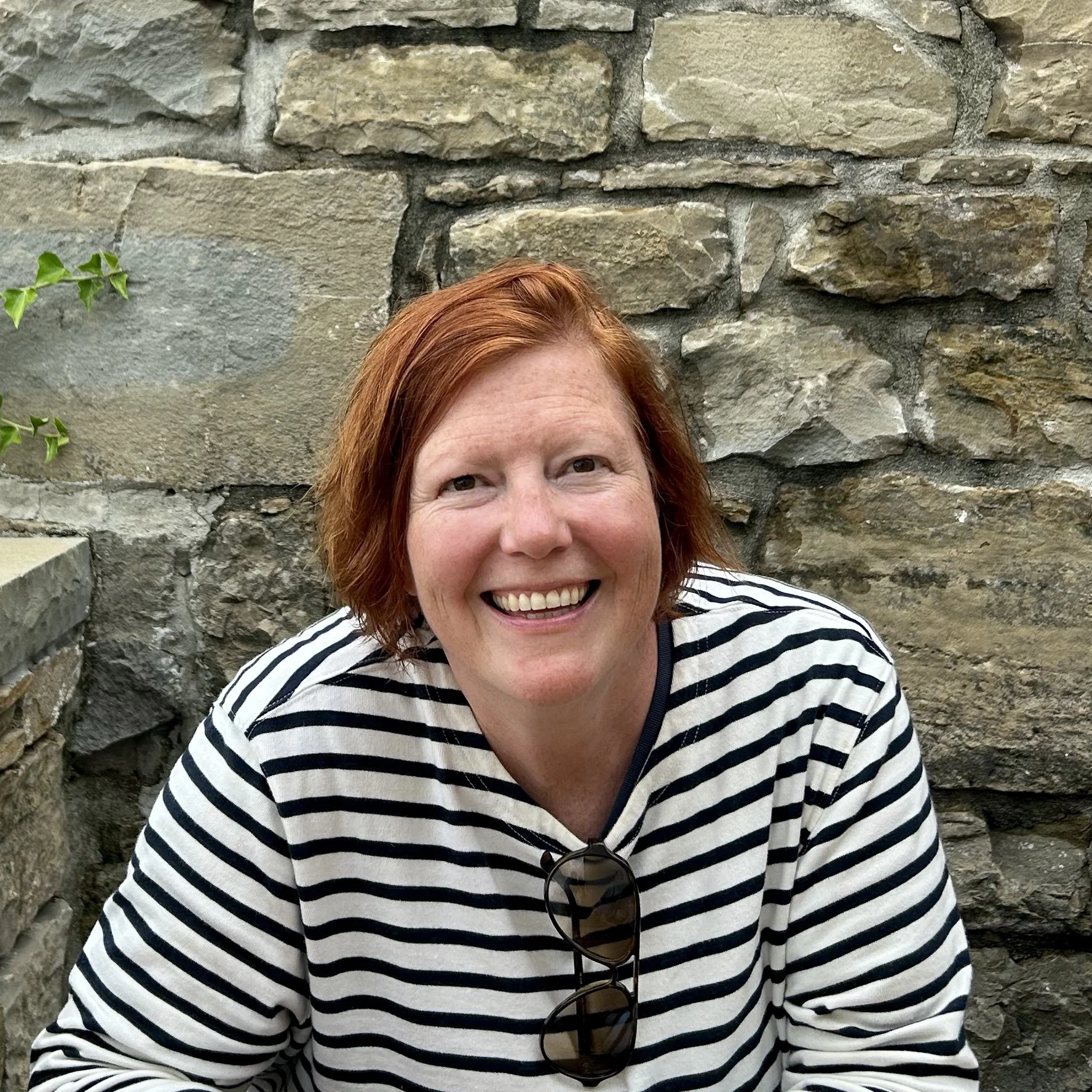 Person smiling in front of a stone wall, wearing a striped shirt and sunglasses.