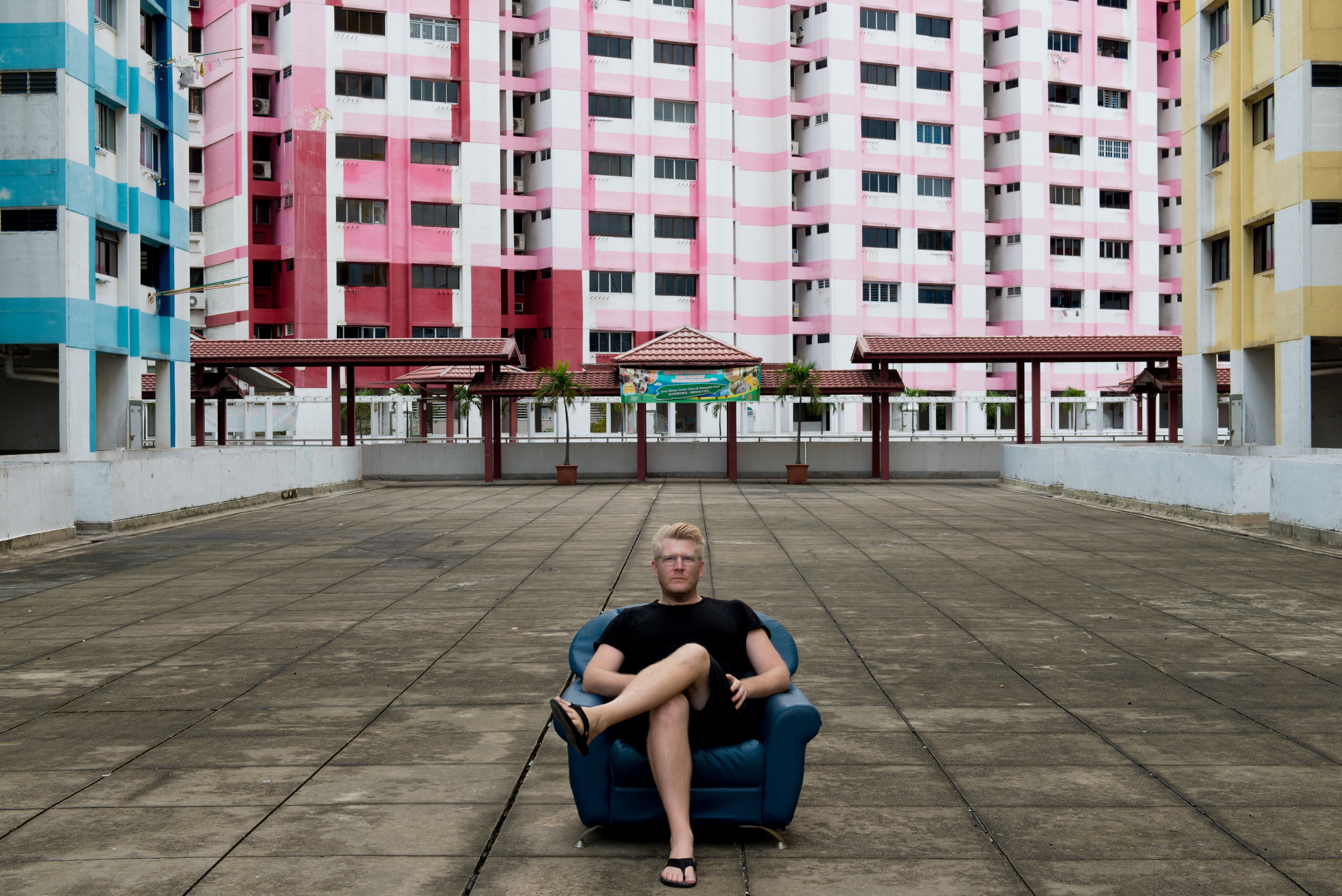 Person sitting in a blue armchair on a concrete terrace with colorful apartment buildings in the background.