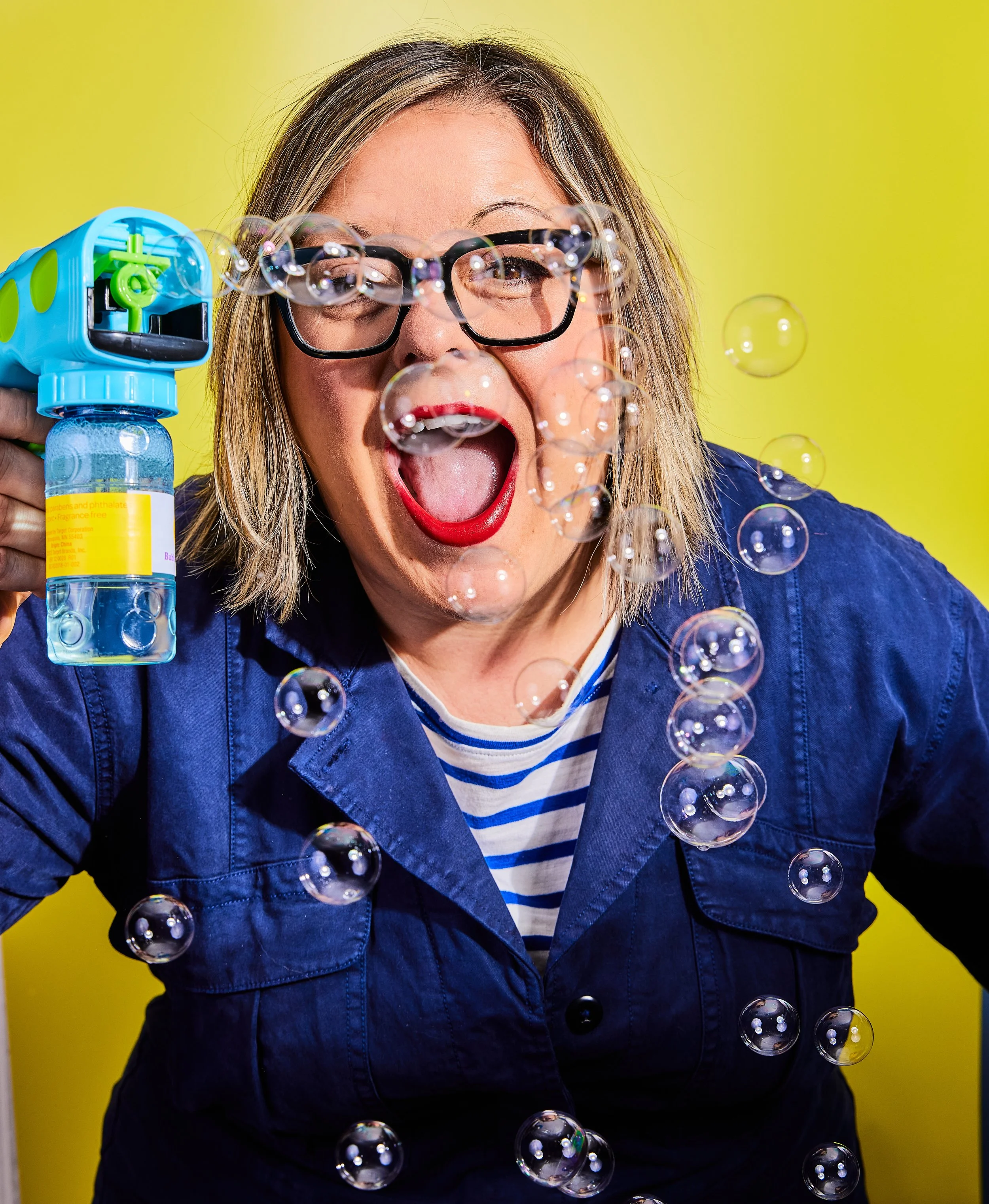 Person with glasses blowing bubbles with a bubble gun against a yellow background.
