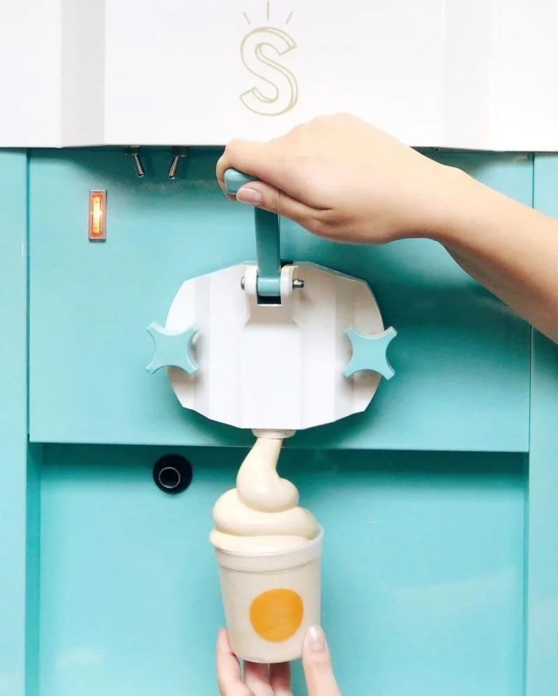 A person's hand dispensing soft serve ice cream from a turquoise machine into a cup.