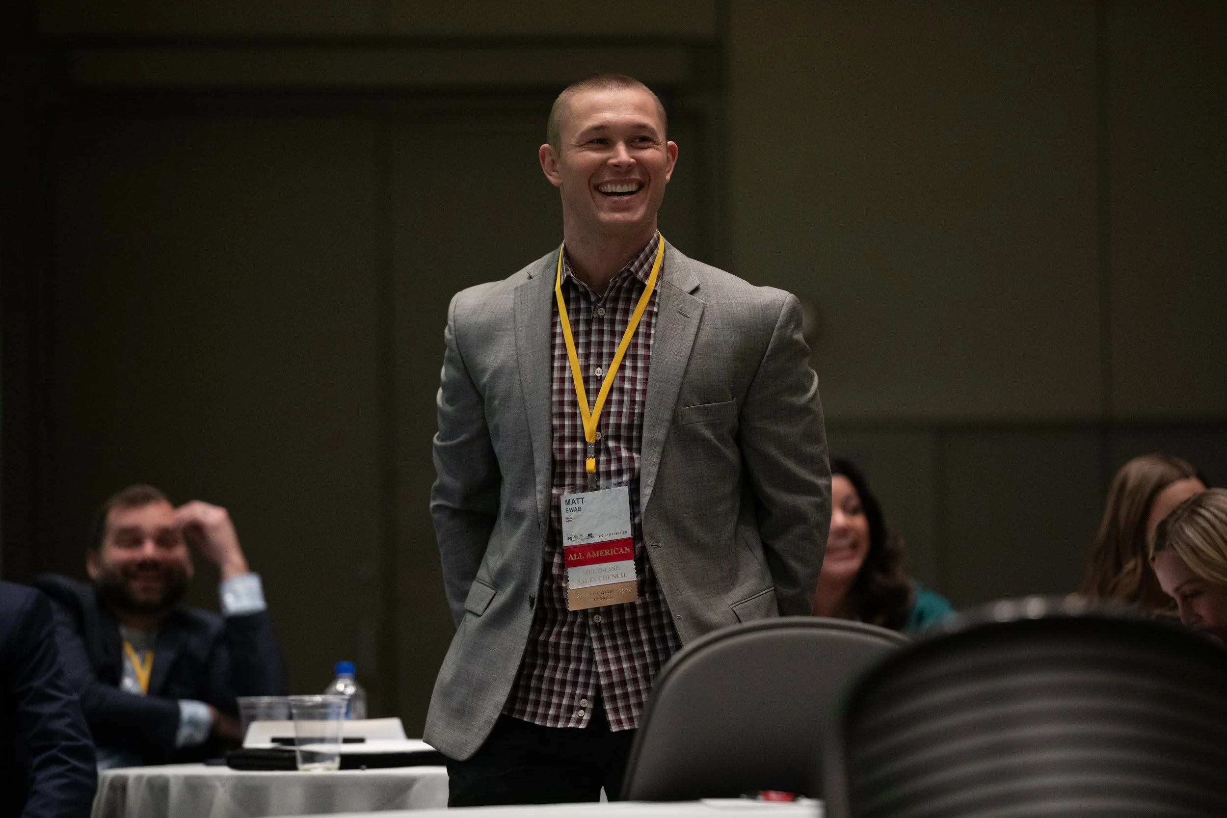 A man in a gray blazer and checkered shirt is smiling at a conference, wearing a yellow lanyard and badge reading "Matt" and "ALL AMERICAN." Others are seated around him.