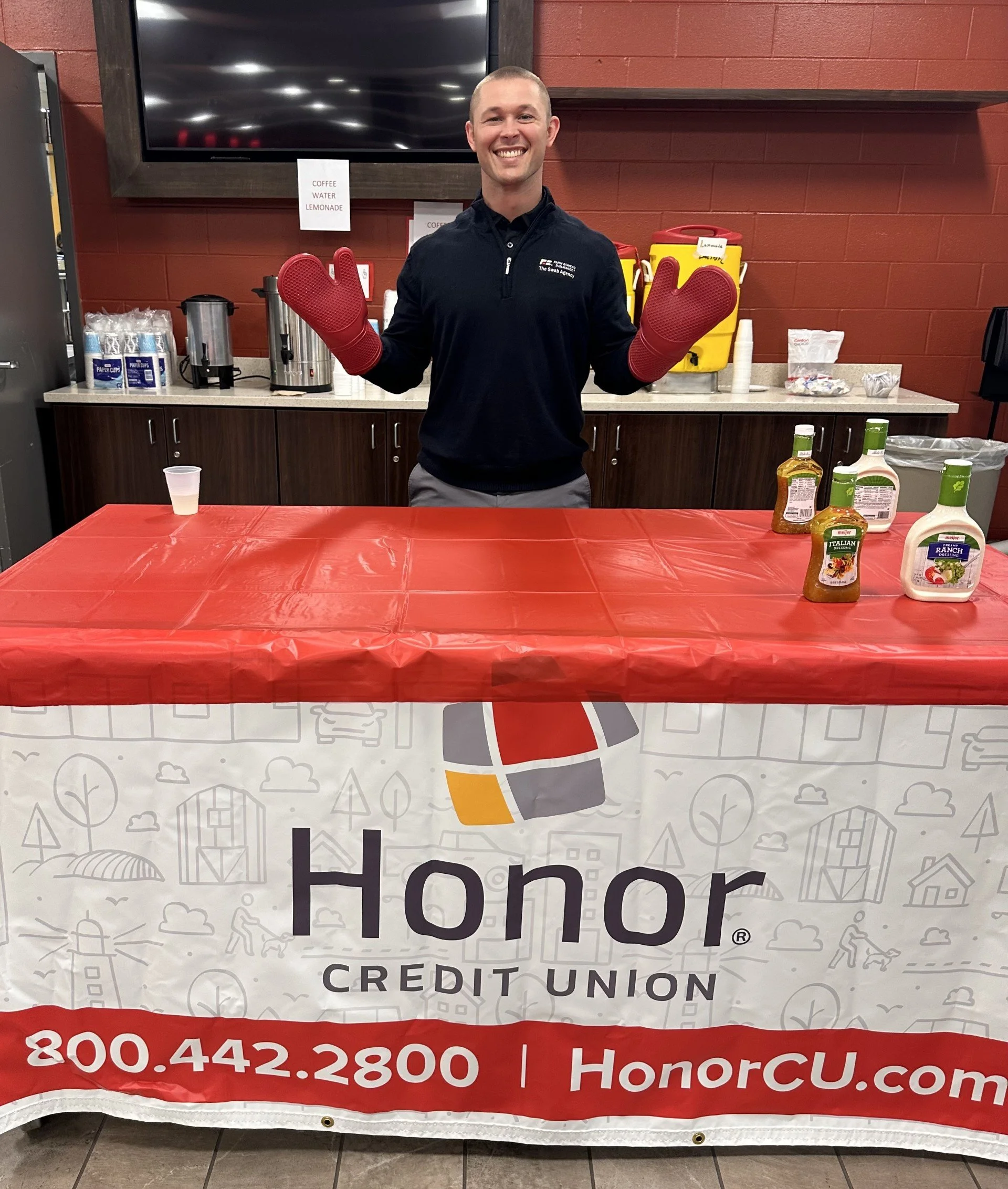 A man smiling and wearing oven mitts standing behind a table with promotional banner for Honor Credit Union. The table has condiments on it, and there is a drink cup on the left. The background includes a coffee station and a large television screen.