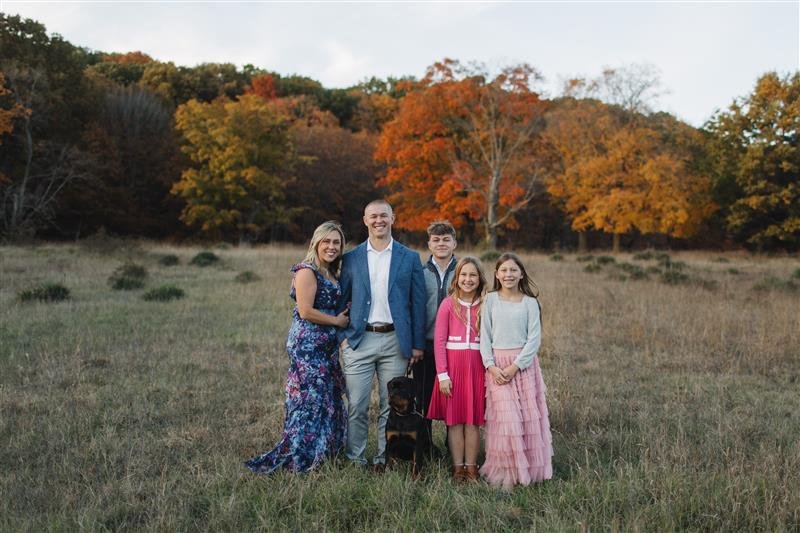 Family of five with a dog standing in a field with colorful fall trees in the background.