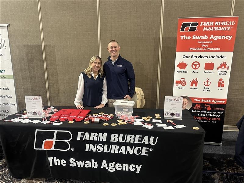 Two representatives standing behind a table promoting Farm Bureau Insurance's The Swab Agency at an event, with a large red and white banner displaying services offered.