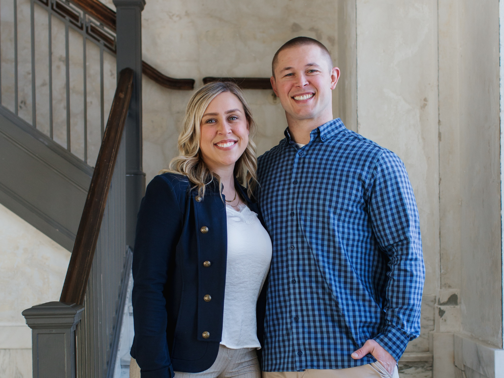A smiling young man and woman standing together indoors, near a staircase, in casual business attire.