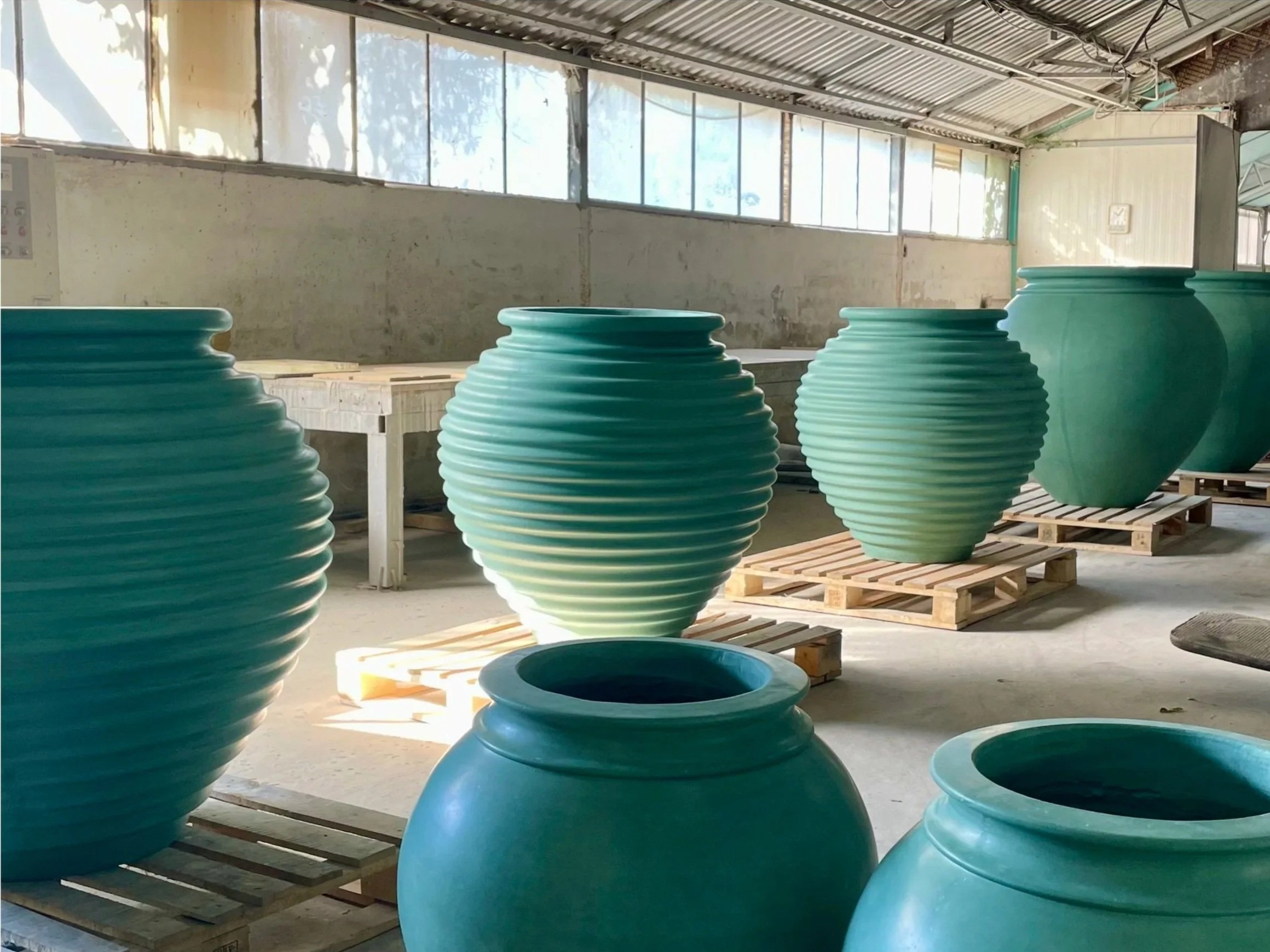 Large turquoise ceramic vases in a warehouse with pallet racks and sunlight pouring through windows.