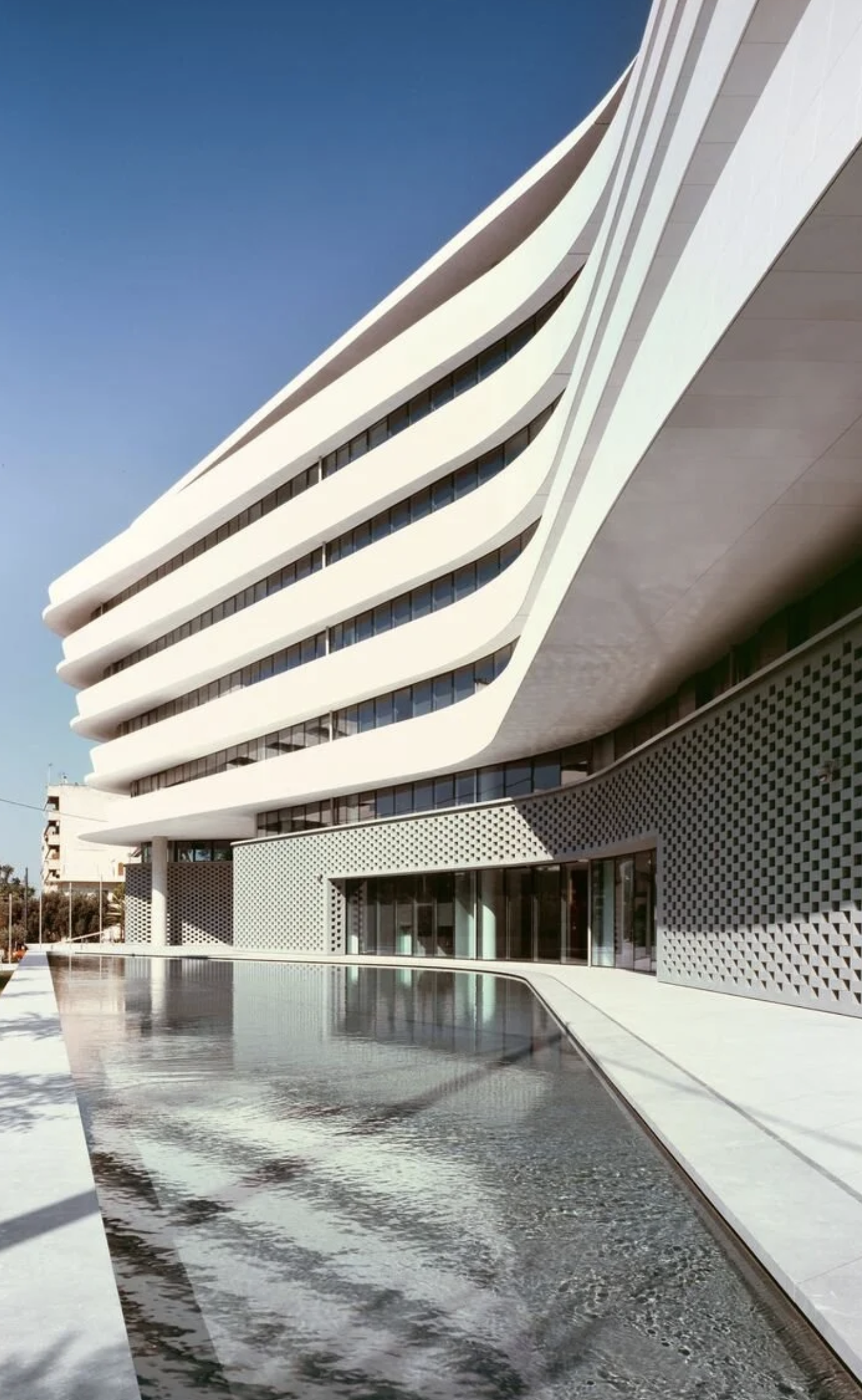 Modern white multi-story building with curved balconies, glass windows, and a reflective water feature in front.