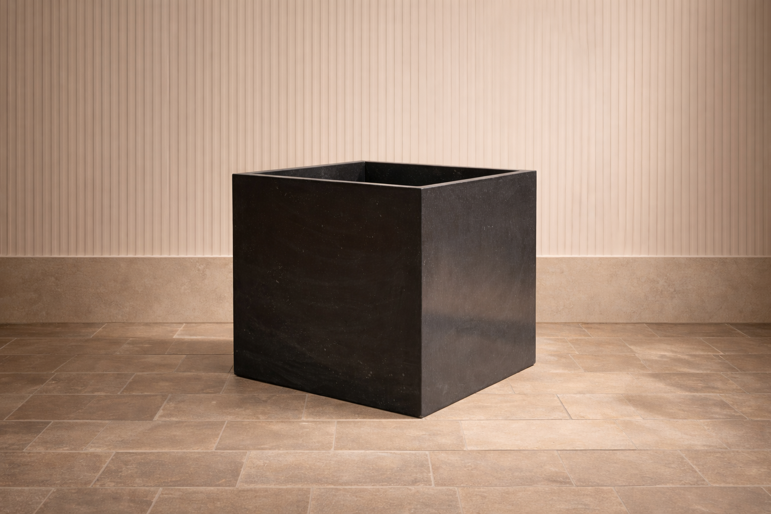 A black square stone planter on a tile floor against a pinkish wall with vertical ridges.