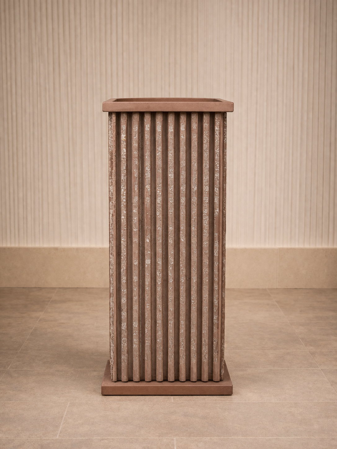 A tall, rectangular, textured brown flower pot with vertical ridges, placed on a tiled floor against a light-colored, vertically striped wall.