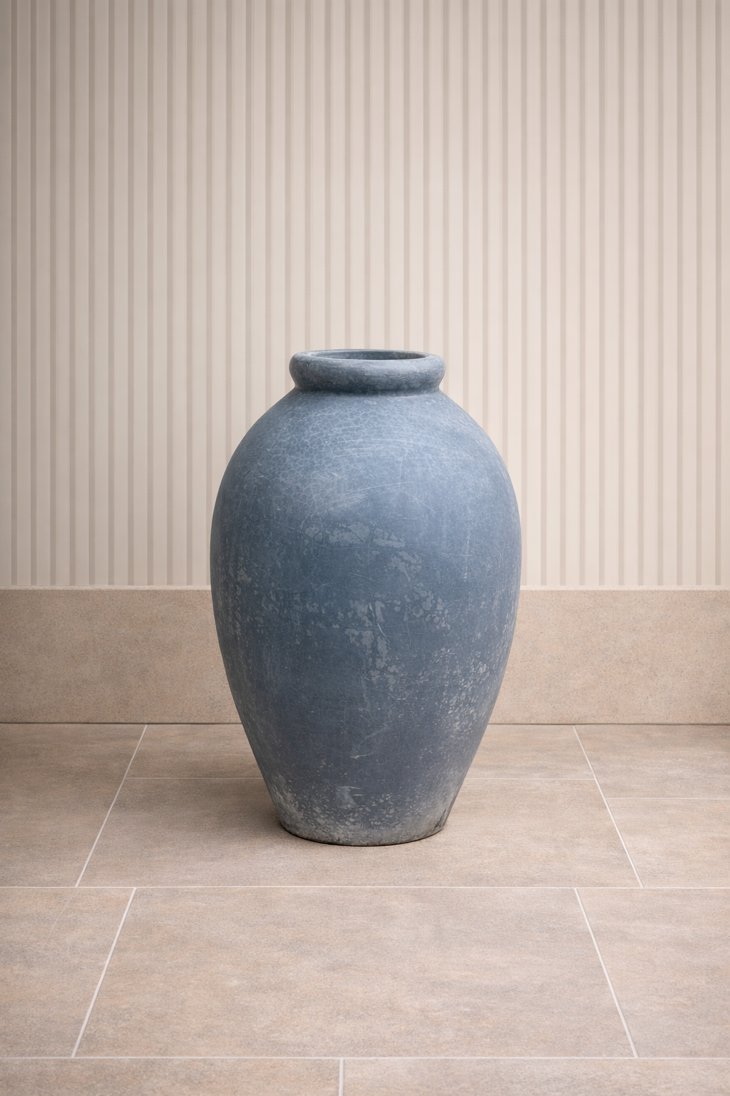 A large blue ceramic vase with a weathered finish, placed on a beige tiled floor against a cream-colored wall with vertical lines.