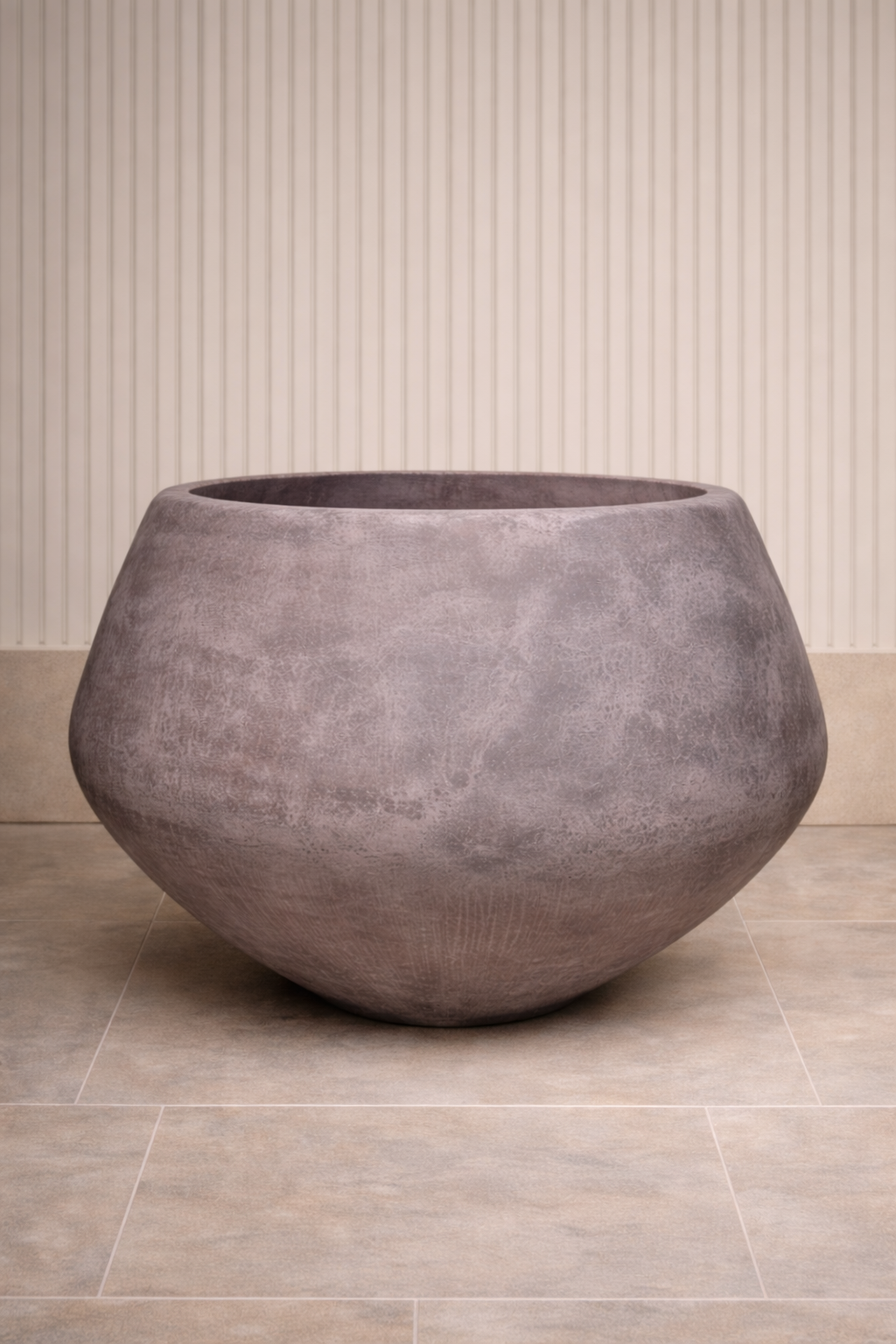 A large, rounded, gray planter with a textured surface, placed on a tiled floor against a wall with vertical striped wallpaper.