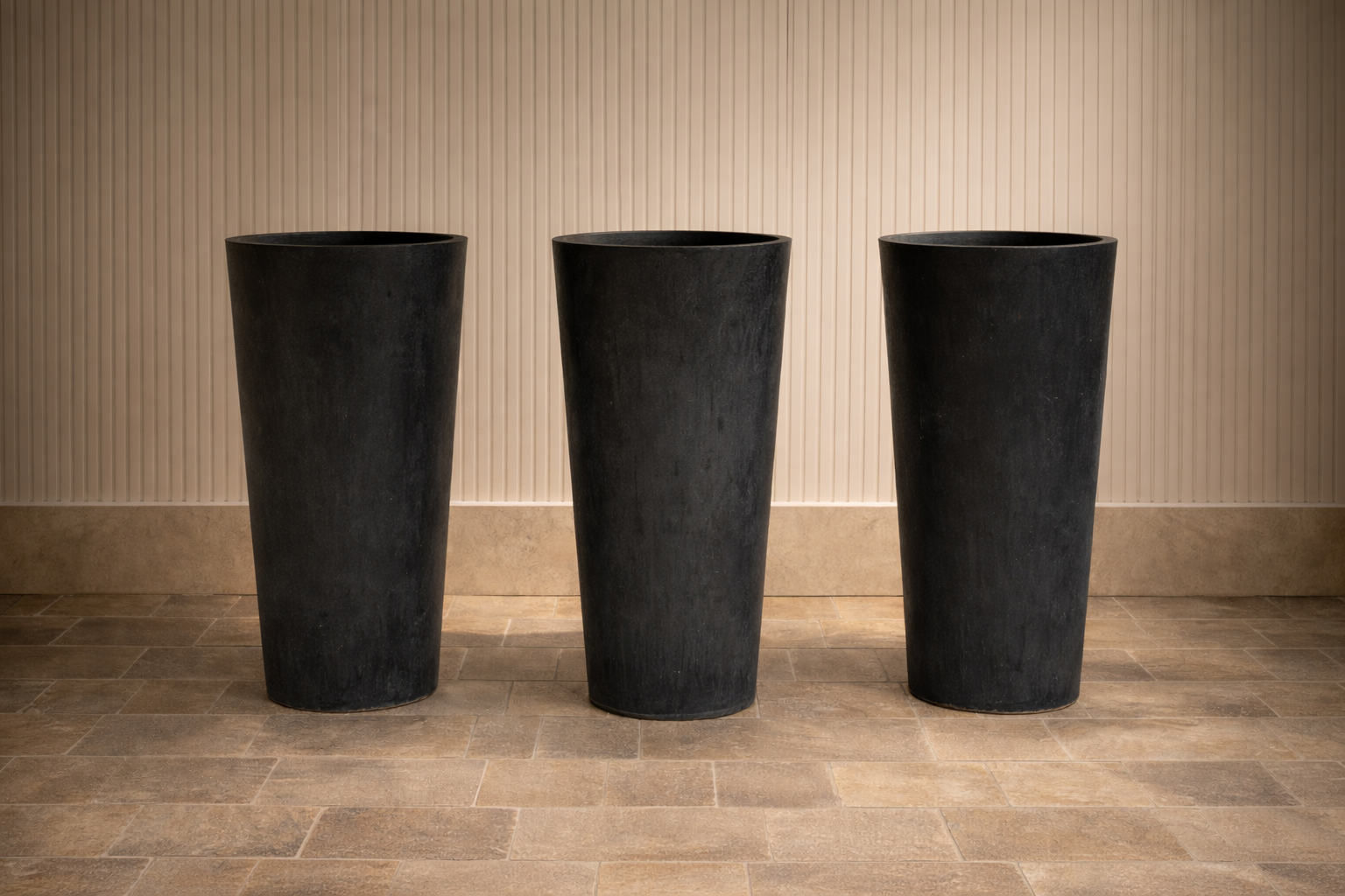 Three large black flowerpots placed on a tiled floor against a beige vertical-striped wall.