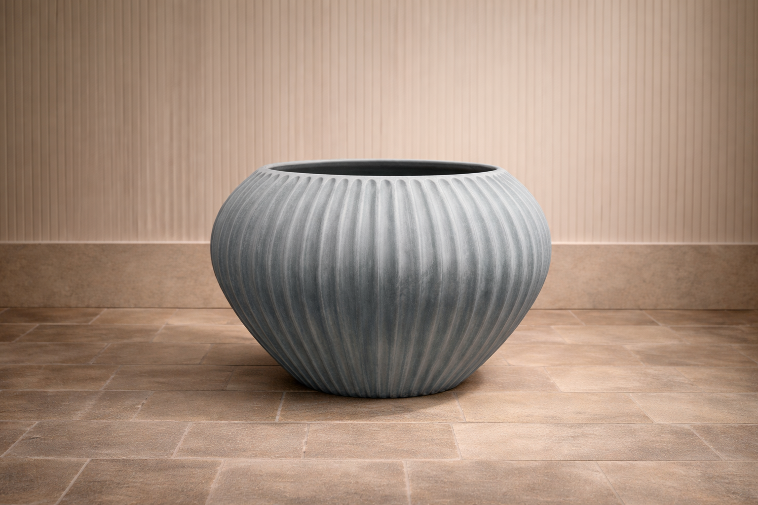 Large round ceramic planter with vertical ribbed design placed on a tiled floor against a beige vertical-lined wall.