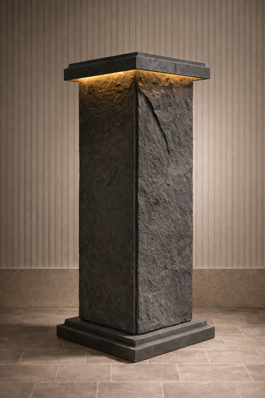 A tall, rectangular, dark stone pedestal with a textured surface, topped with a smooth, flat stone cap, with warm lighting highlighting the edges. It is placed against a vertically striped wall on a tiled floor.