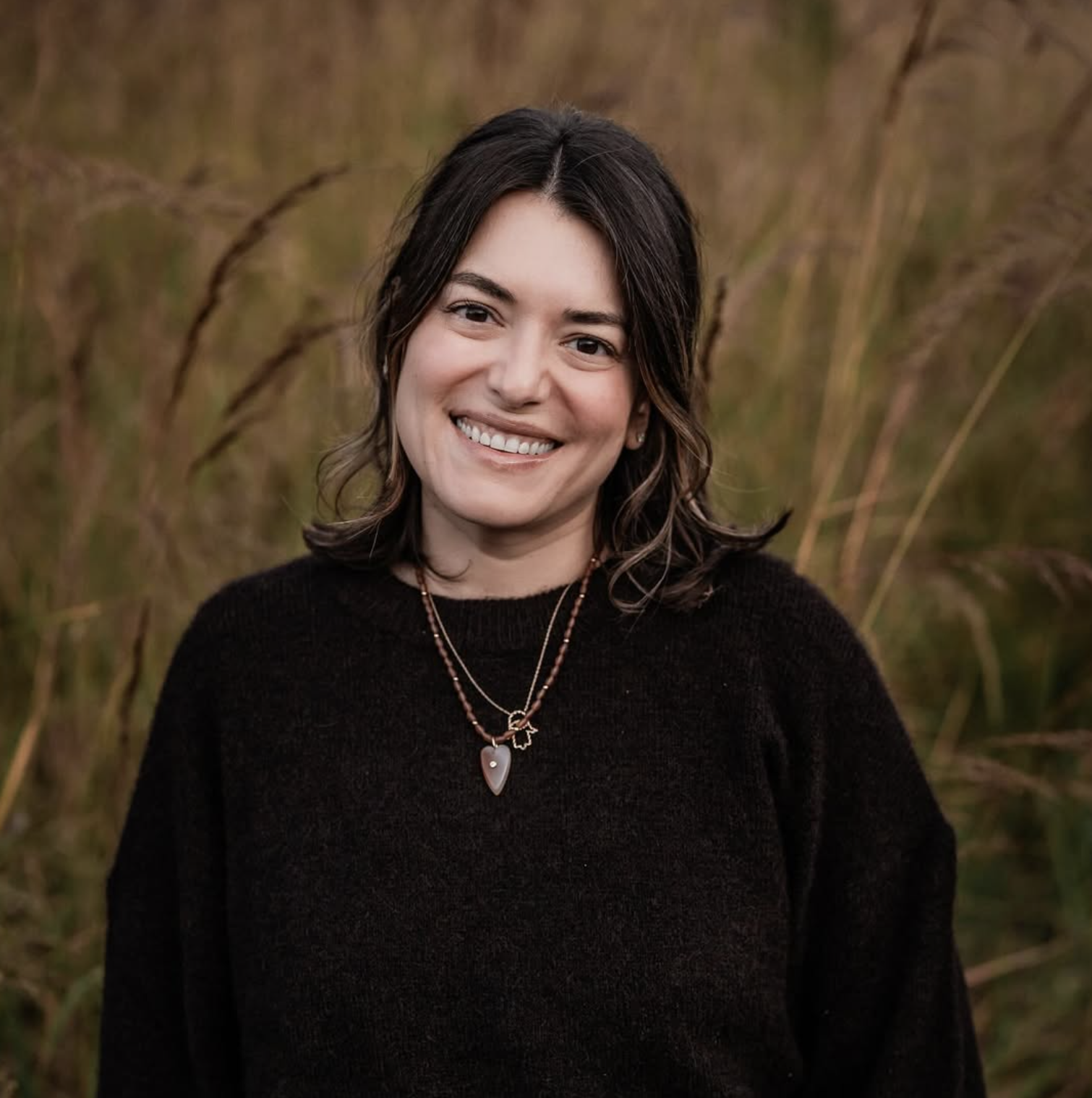 A woman with shoulder-length dark hair, smiling, wearing a black sweater and layered necklaces, standing outdoors in a field with tall grasses.