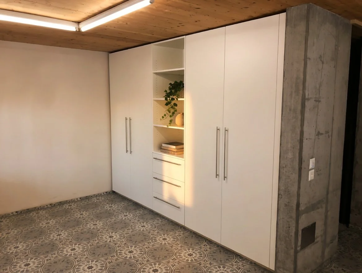 Weiße Einbauschränke mit langen Griffen neben einer Wand aus rohem Beton, indirekte Beleuchtung um die Schränke, gepflanzte Dekoration und Bücher auf offenen Regalbulten, geradliniges modernes Design.