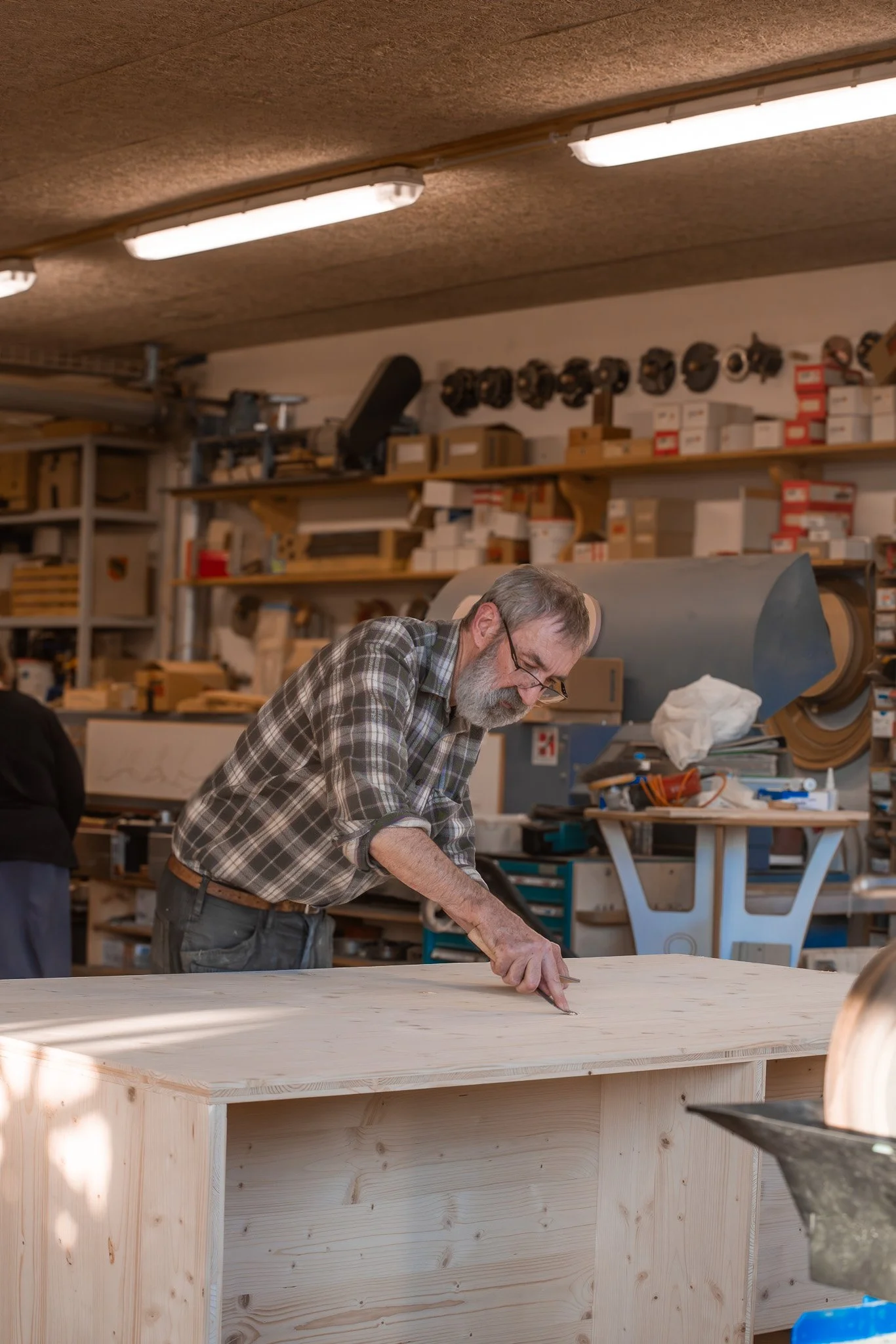 Ein älterer Mann arbeitet in einer Werkstatt an einem großen Holztisch, umgeben von Werkzeugen und Materialien.