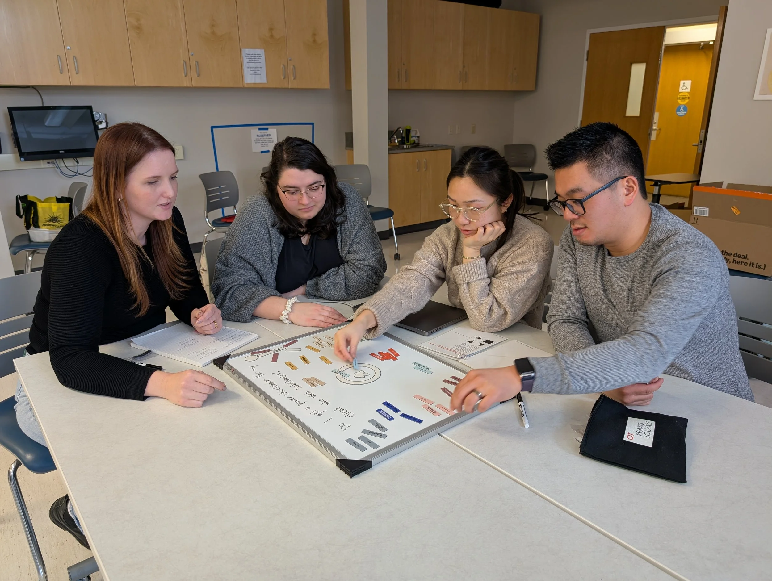 Five diverse people sitting at a table in a conference room, engaging in a discussion around a whiteboard with colorful sticky notes and diagrams.