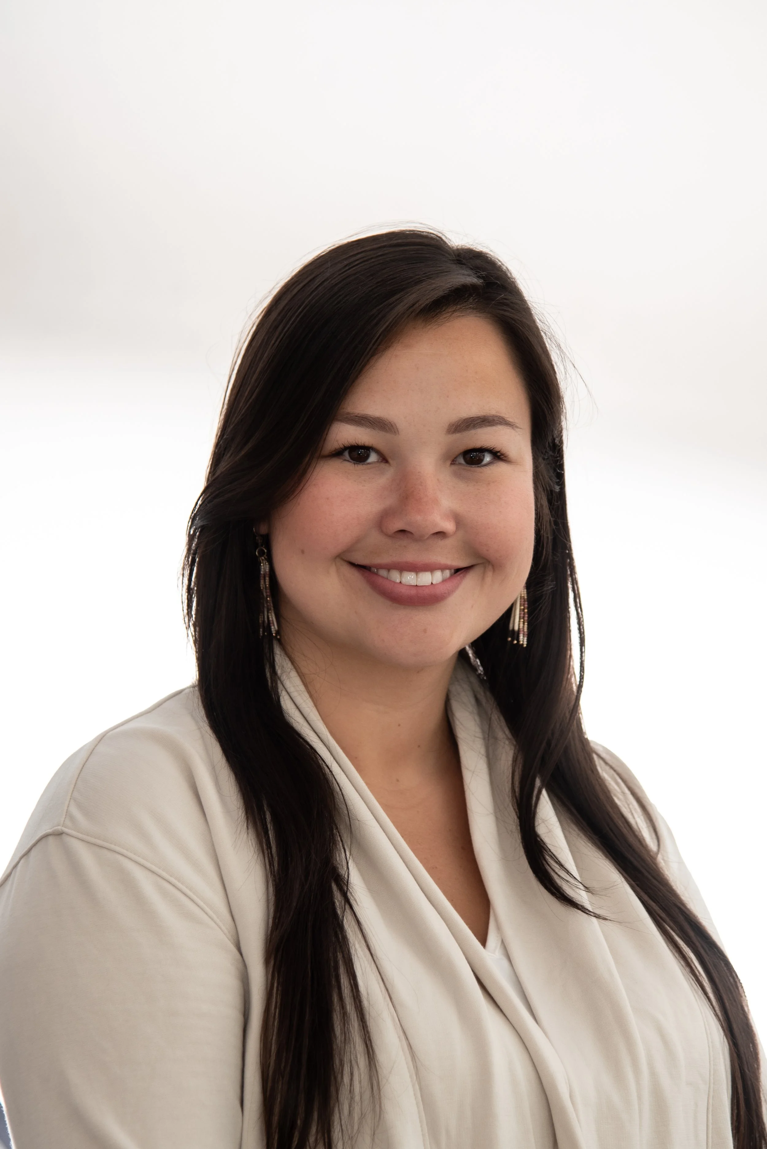 Portrait of a young woman with long dark hair, smiling, wearing a cream-colored top and earrings, against a white background.