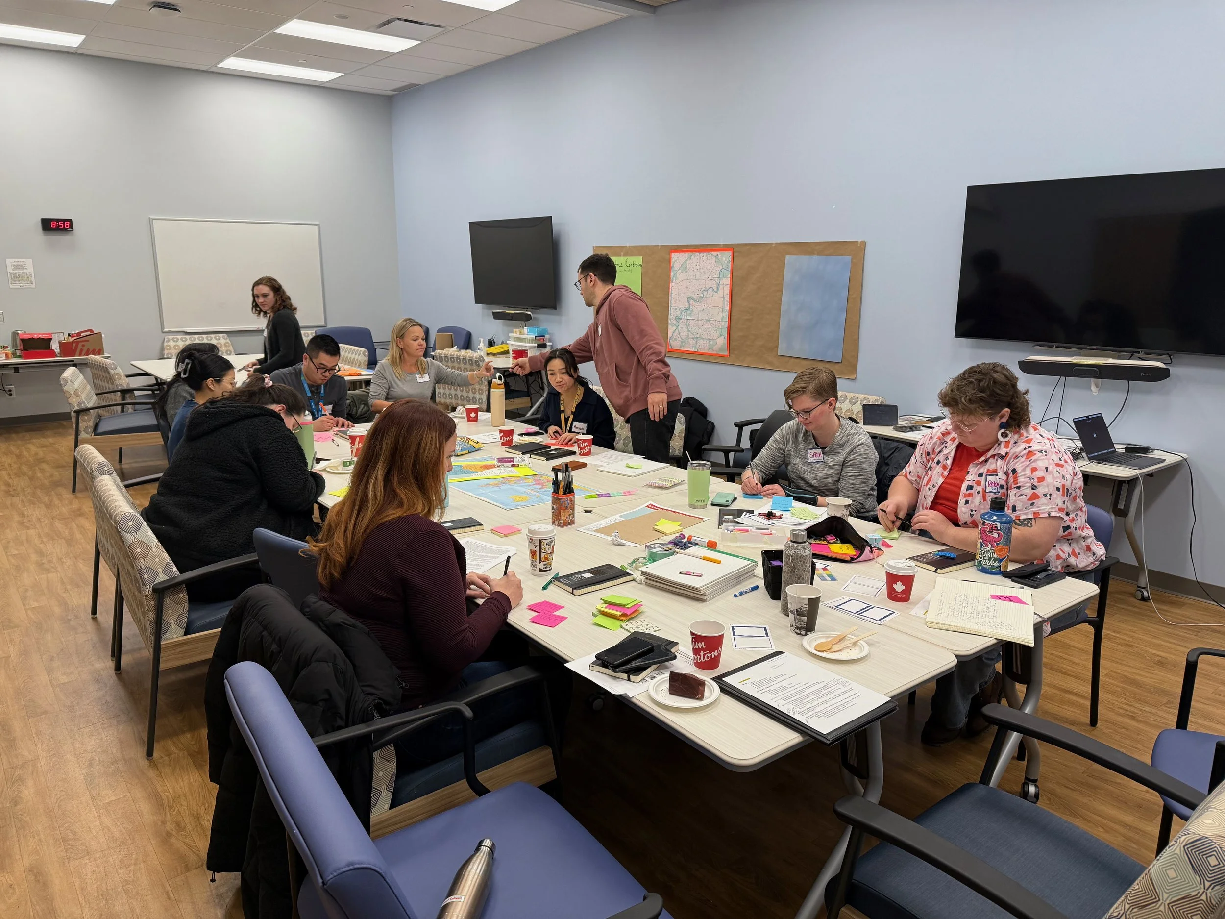 A group of people gathered around a large conference table in a meeting room, engaged in a workshop or collaborative activity, with various supplies, papers, and snacks on the table.
