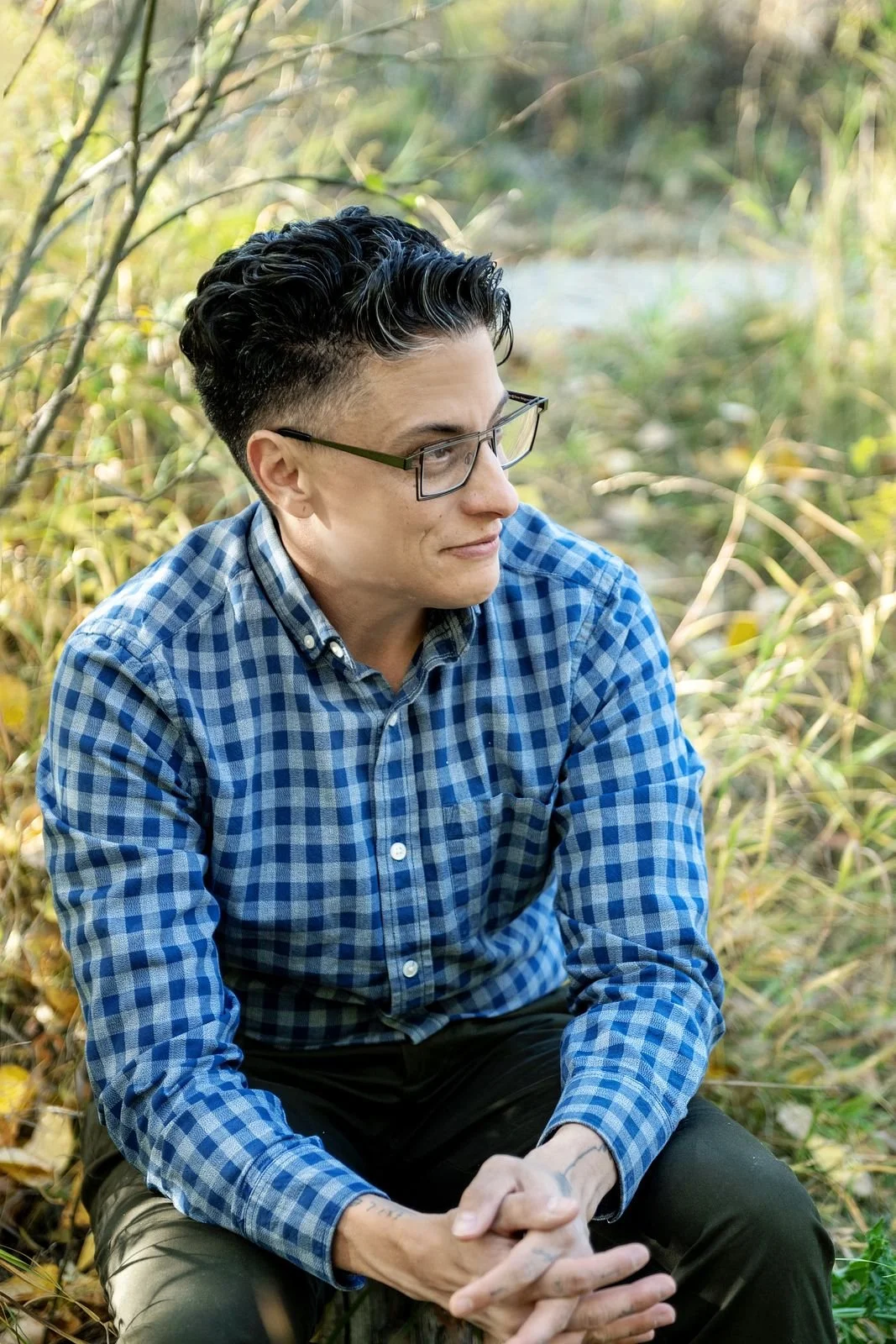 A person with glasses and short dark hair, wearing a blue checkered shirt, sitting outdoors in a natural setting with green foliage and grass in the background.