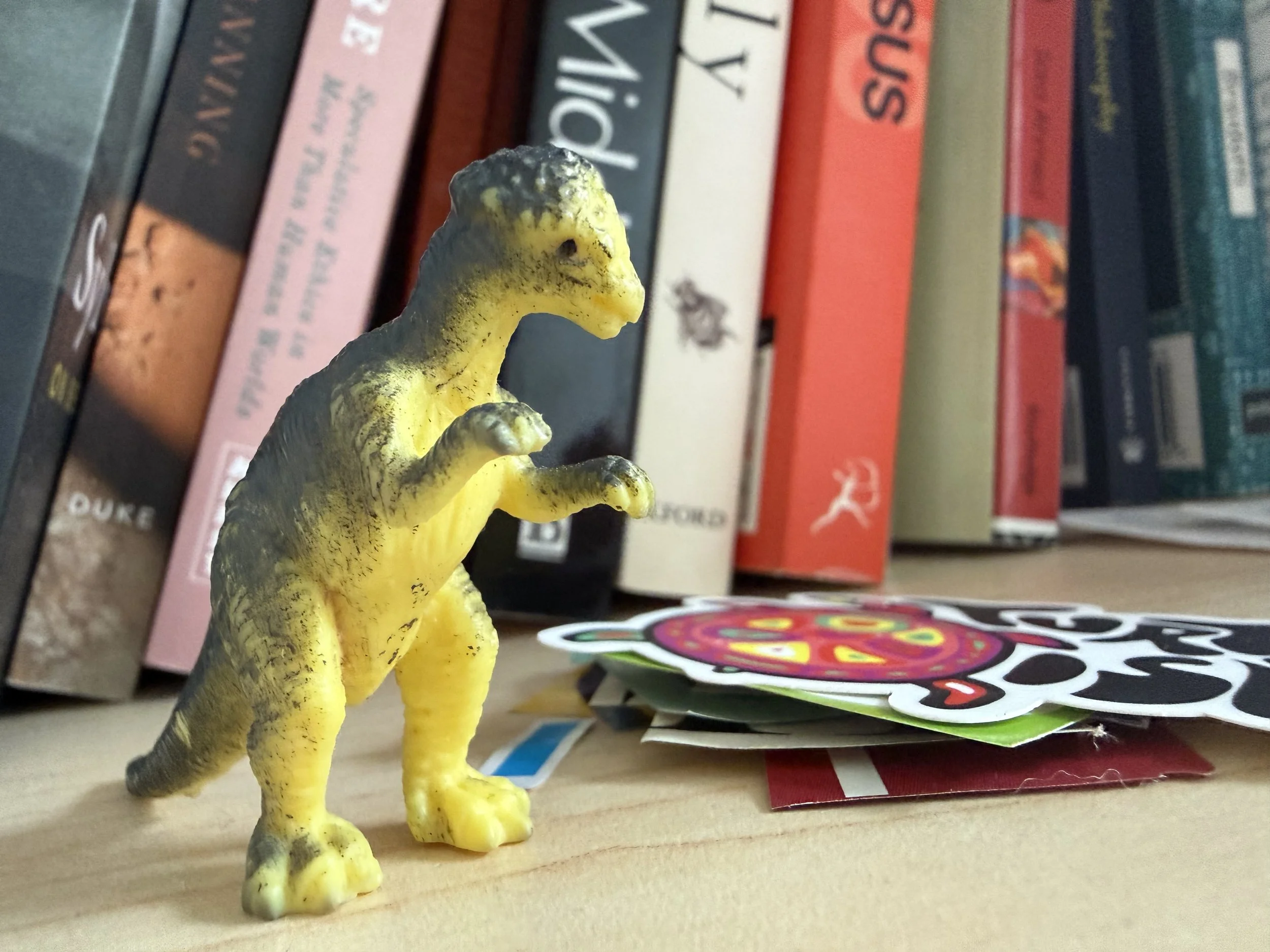 A small plastic yellow dinosaur figurine with black spots standing on a wooden surface in front of a row of books and some stickers.
