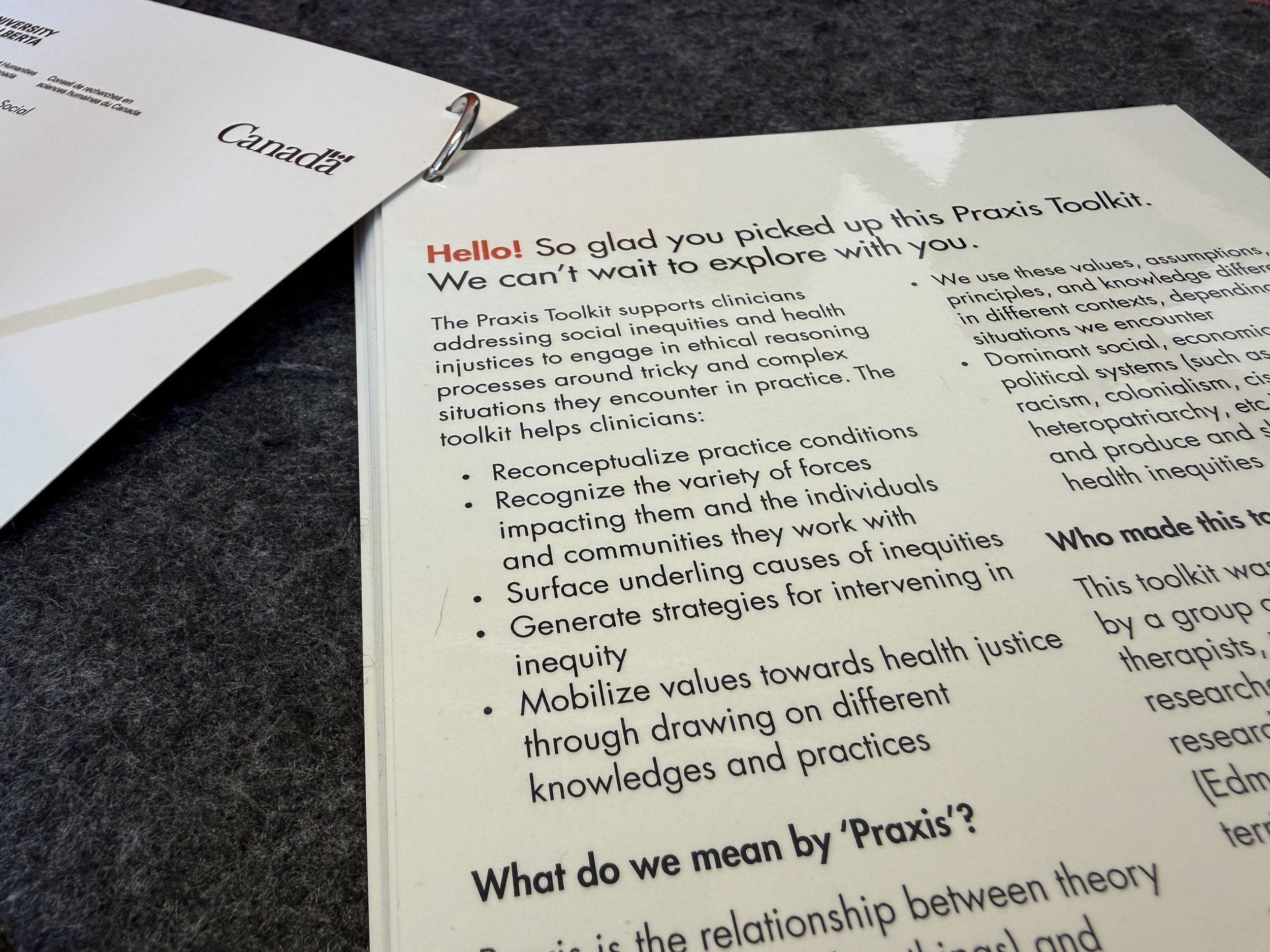 Close-up of a printed document titled 'Praxis Toolkit' with a heading in red 'Hello!' and text supporting social equity and health justice, laid on a dark surface. Partially visible paper on the left has the word 'Canada'.