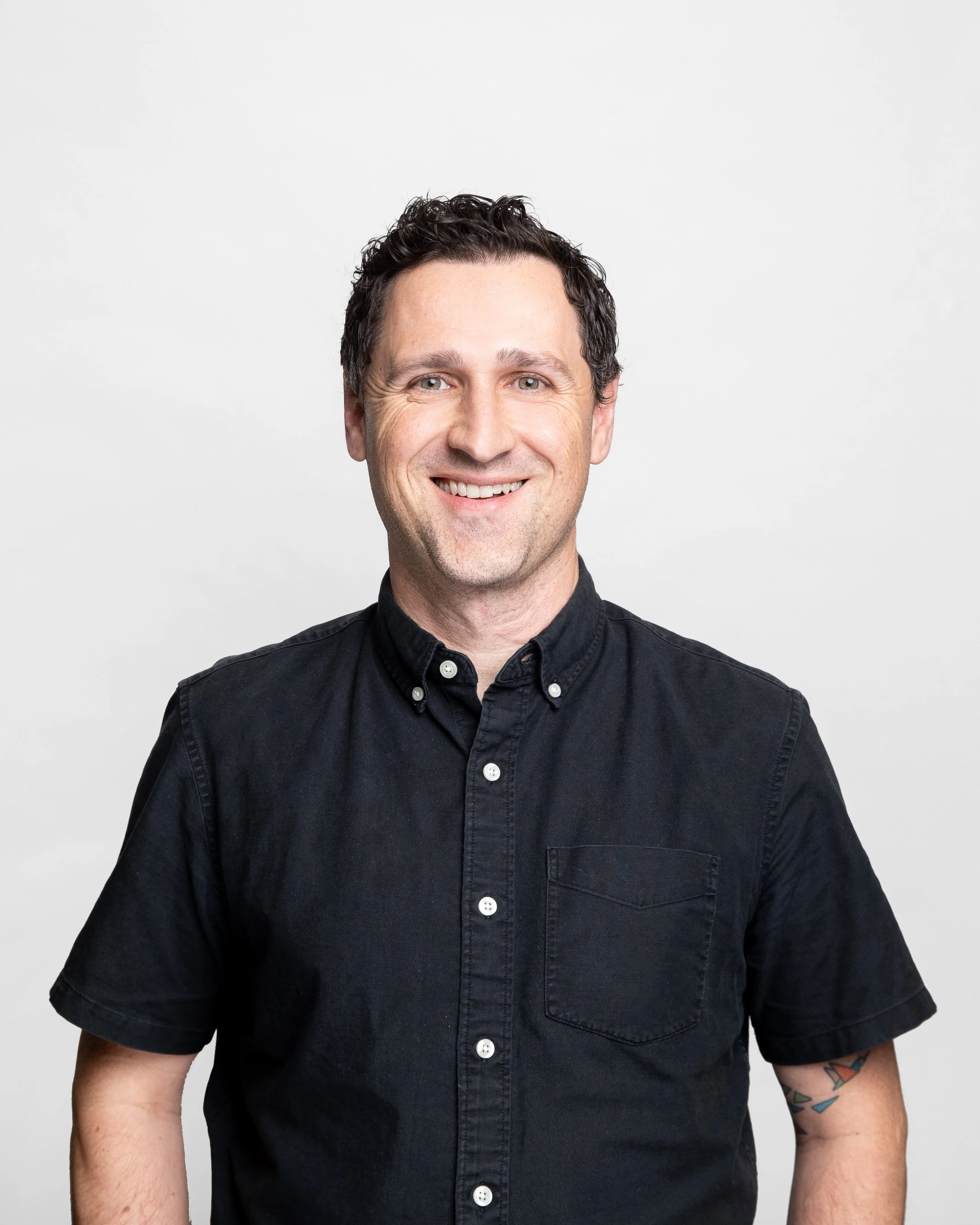 A smiling man with dark, curly hair, wearing a black button-up shirt, posing against a plain light gray background.