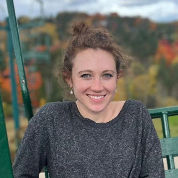 A woman with curly hair tied up in a bun, smiling, wearing a dark gray sweater, sitting outdoors on a green metal structure with a blurred autumn landscape in the background.
