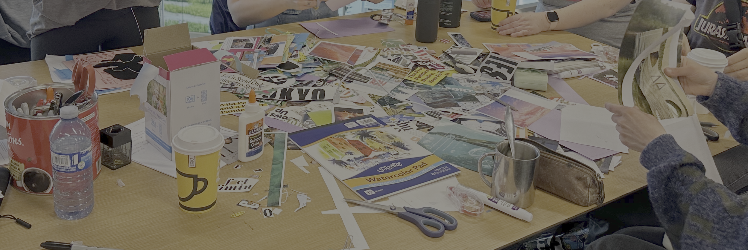 A cluttered table with art supplies including scissors, glue, markers, a watercolor pad, magazines, a water bottle, and paper. Several people are working with magazines and paper cutouts.
