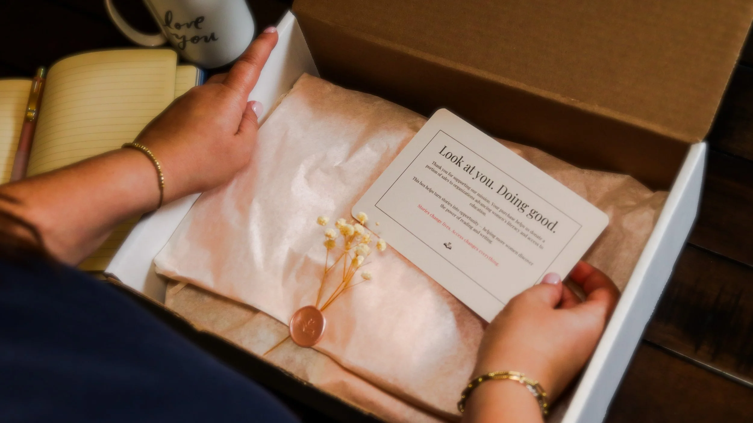 Person holding an open box with pink tissue paper, a small card, and a sprig of white flowers with a wax seal. A mug with 'love you' written on it is in the background.