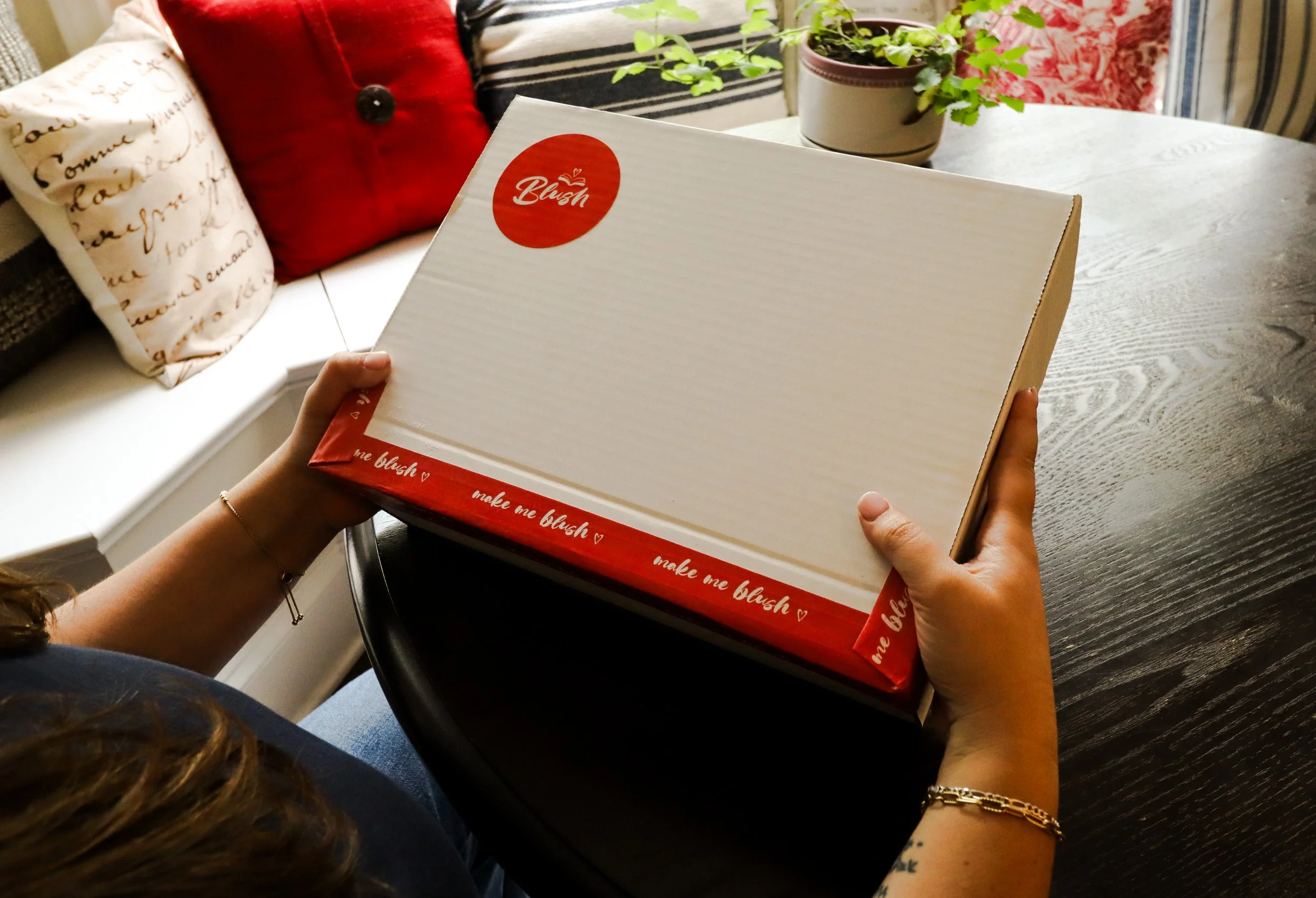 Person holding a white box with red accents labeled 'Blush' and the phrase 'make me blush' on the border, sitting at a dark wooden table.