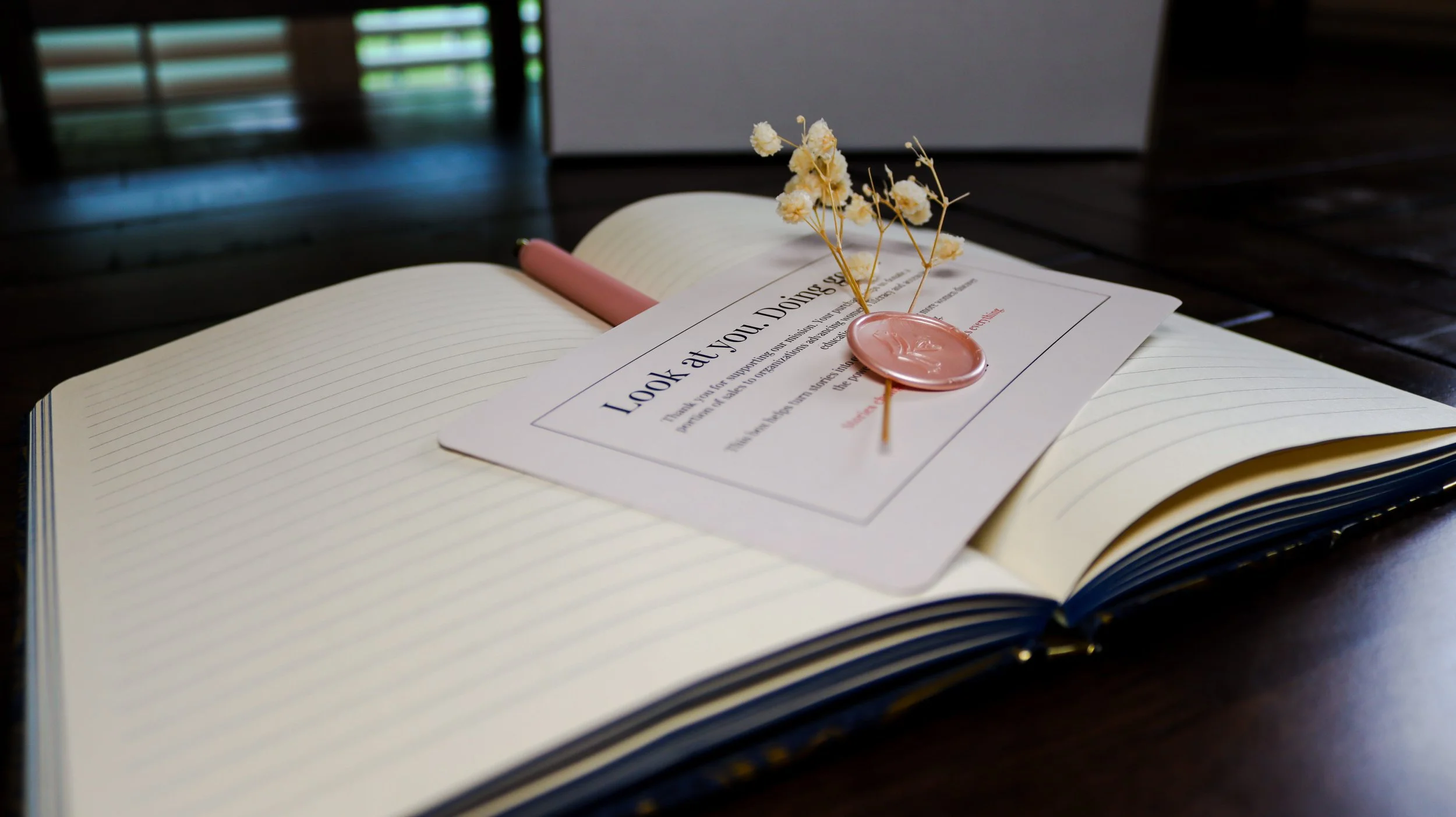 Open notebook with blank lined pages on a dark wooden table, with a pink pen resting on its spine. On top of the notebook is a white cardand a pink wax seal with a small white dried flower resting on it. Background shows a window with blinds letting in natural light.