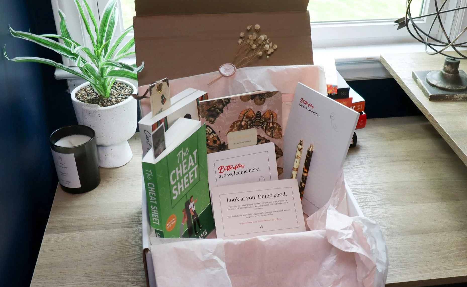 A gift basket with books, stationery, and decorative items on a wooden table near a window, with a potted plant and candle nearby.