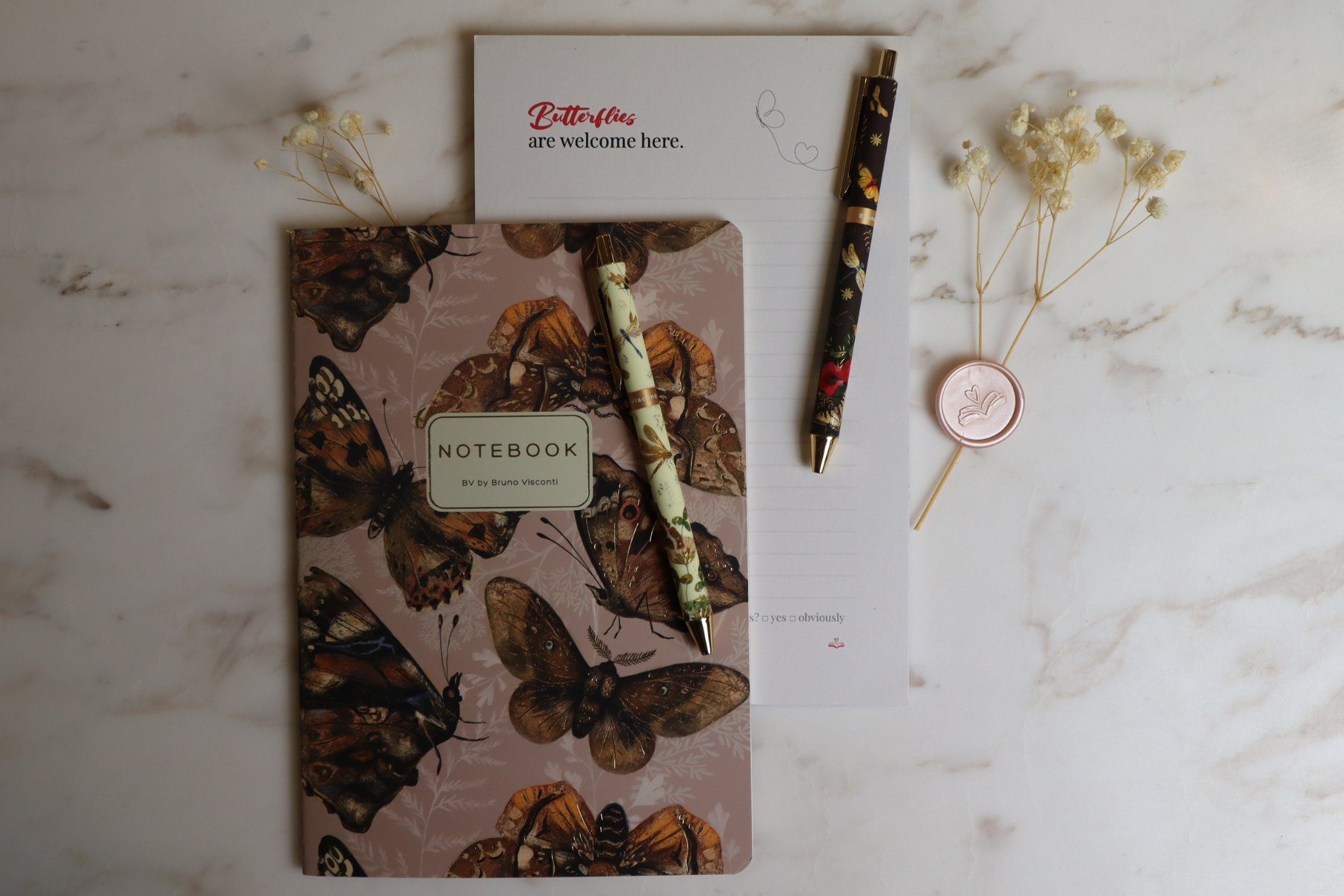A pink butterfly-themed notebook with two pens resting on it, a white piece of paper with text and a butterfly doodle, a few sprigs of dried flowers, and a pink wax seal with floral design on a white marble surface.