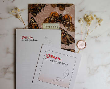 Two notebooks with butterfly and floral covers, and two notepad cards with the message "Butterflies are welcome here," on a white marble surface, accompanied by small dried flowers.