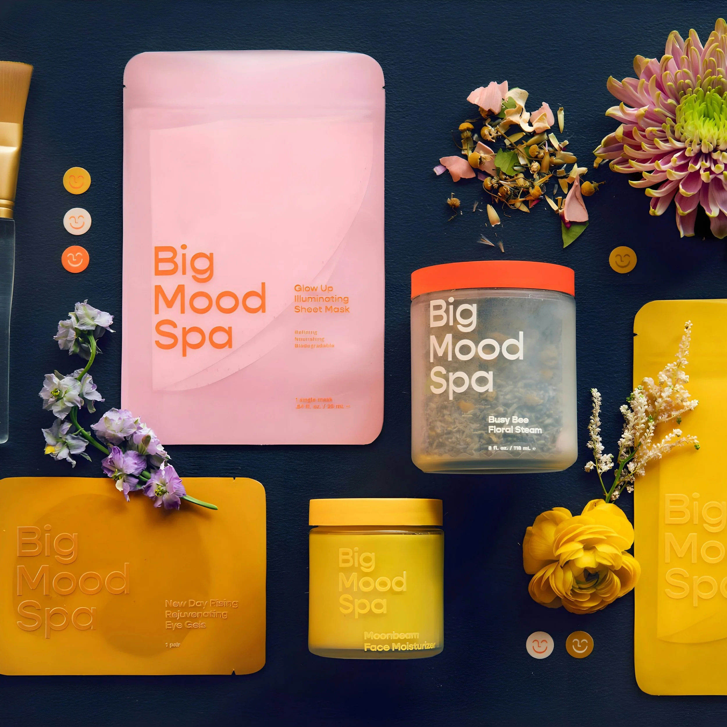 Assorted skincare products from Big Mood Spa including a pink glow-up illuminating sheet mask, a floral steam container, a face moisturizer jar, and two yellow treatment masks, decorated with flowers and small smiley face stickers on a dark background.