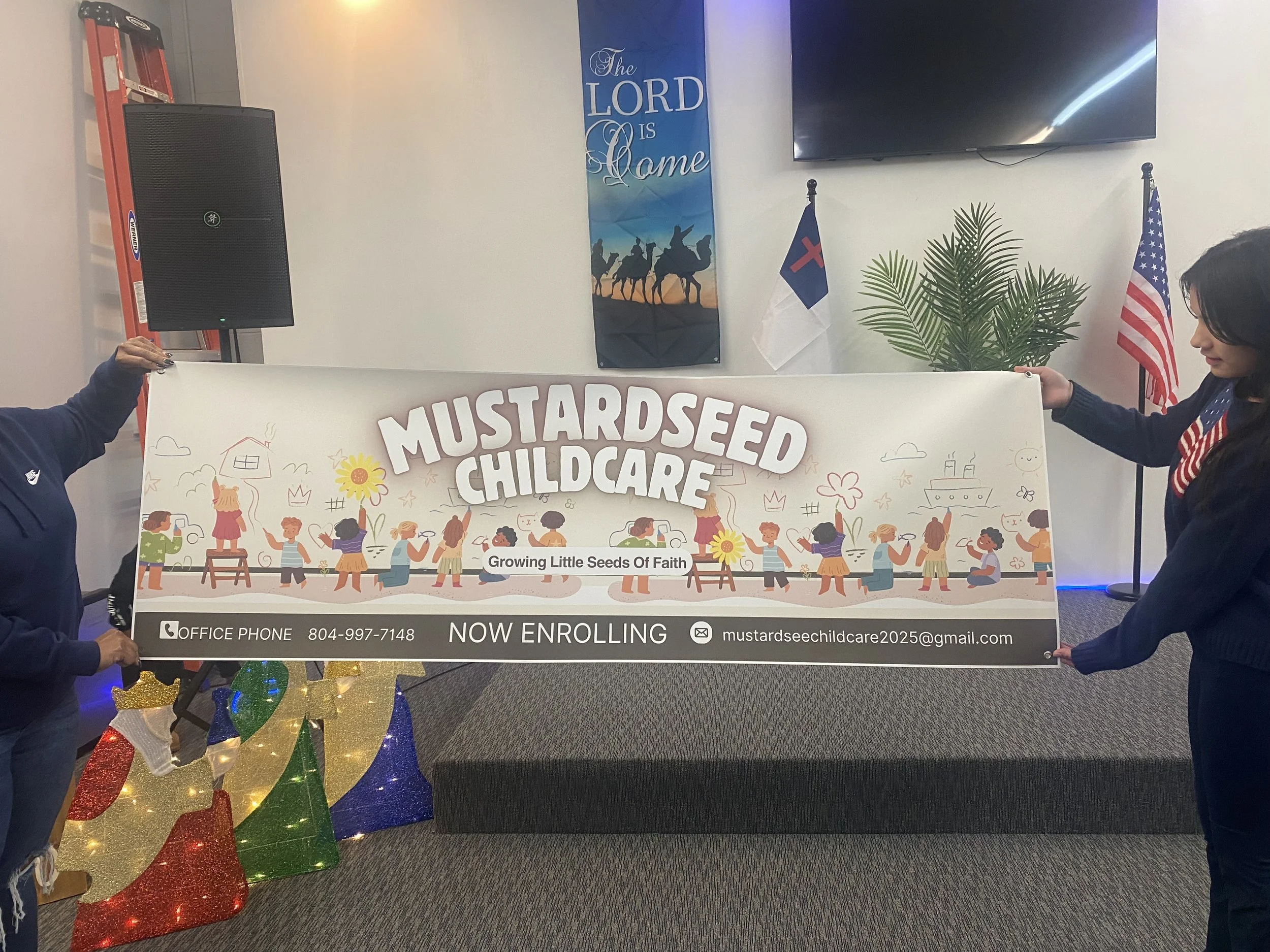 Two women hold a large banner advertising Mustardseed Childcare, with information about enrollment, contact email, and phone number, in a decorated room with American and Christian flags, a large plant, a wall-mounted TV, a poster with a biblical verse, and Christmas decorations.