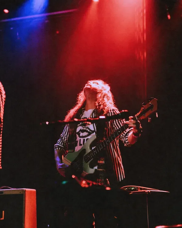 A person with long, curly hair playing an electric guitar on stage under red and blue lights.