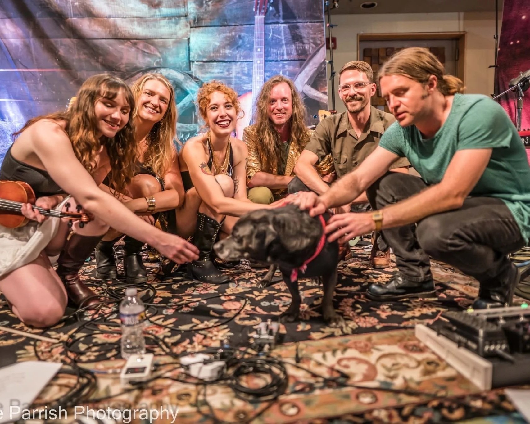 A group of six people with a dog on a colorful carpeted stage, smiling and petting the dog, with musical equipment and a colorful backdrop with a guitar in the background.
