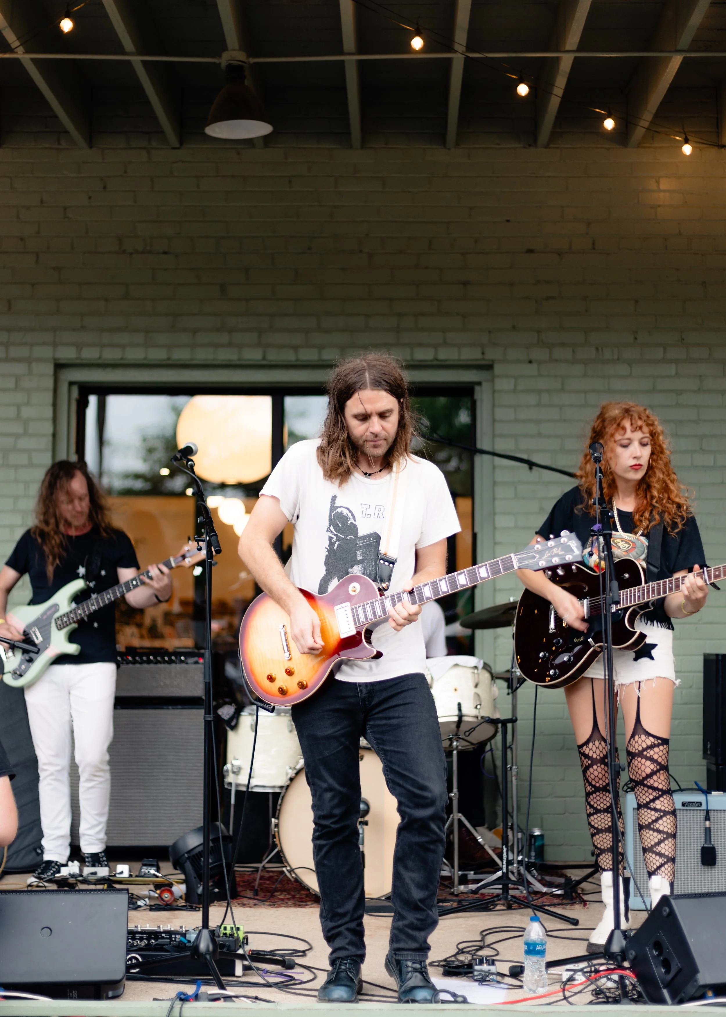A band performing live on an outdoor stage with three guitarists, a drummer, and microphones, against a brick wall with window and string lights overhead.