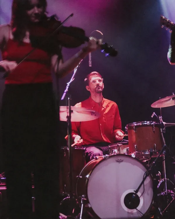 A female musician playing a violin and a male musician playing drums on a stage, with purple lighting.